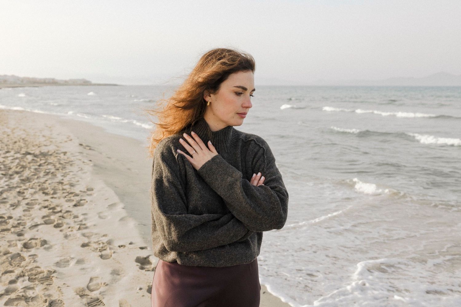 A person in a dark sweater stands on a windy beach with waves crashing behind them on an overcast day.