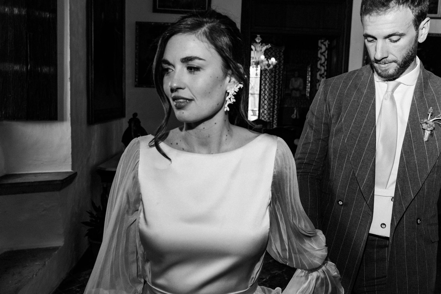 A black and white photo of a bride in a sleek dress with sheer sleeves walking alongside someone in a suit.
