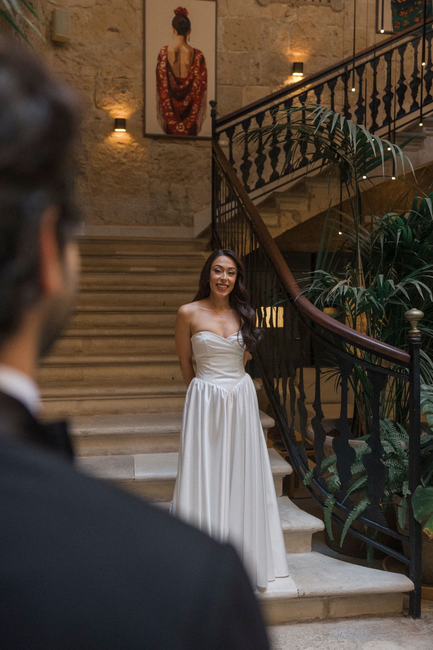 A bride in a flowing white dress stands gracefully on an elegant stone staircase with ornate railings and warm lighting.