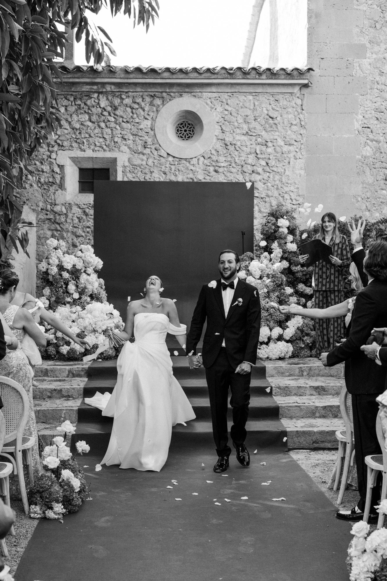 Wedding party sequence captured against stone wall with white flowers and green floor.