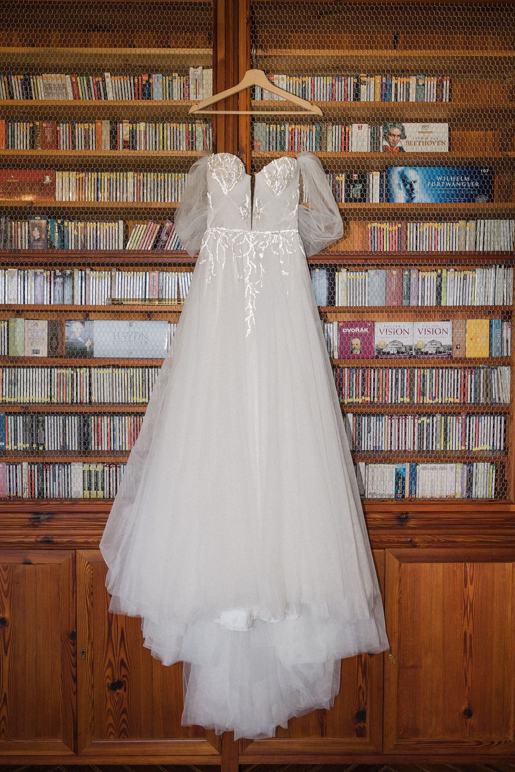White tulle wedding dress hangs in front of wooden library shelves filled with books.
