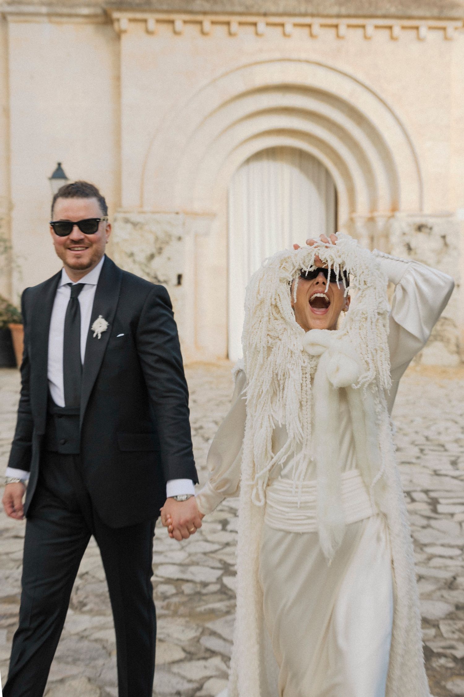 Joyful wedding couple posing in front of an arched doorway with bride wearing fringe details.