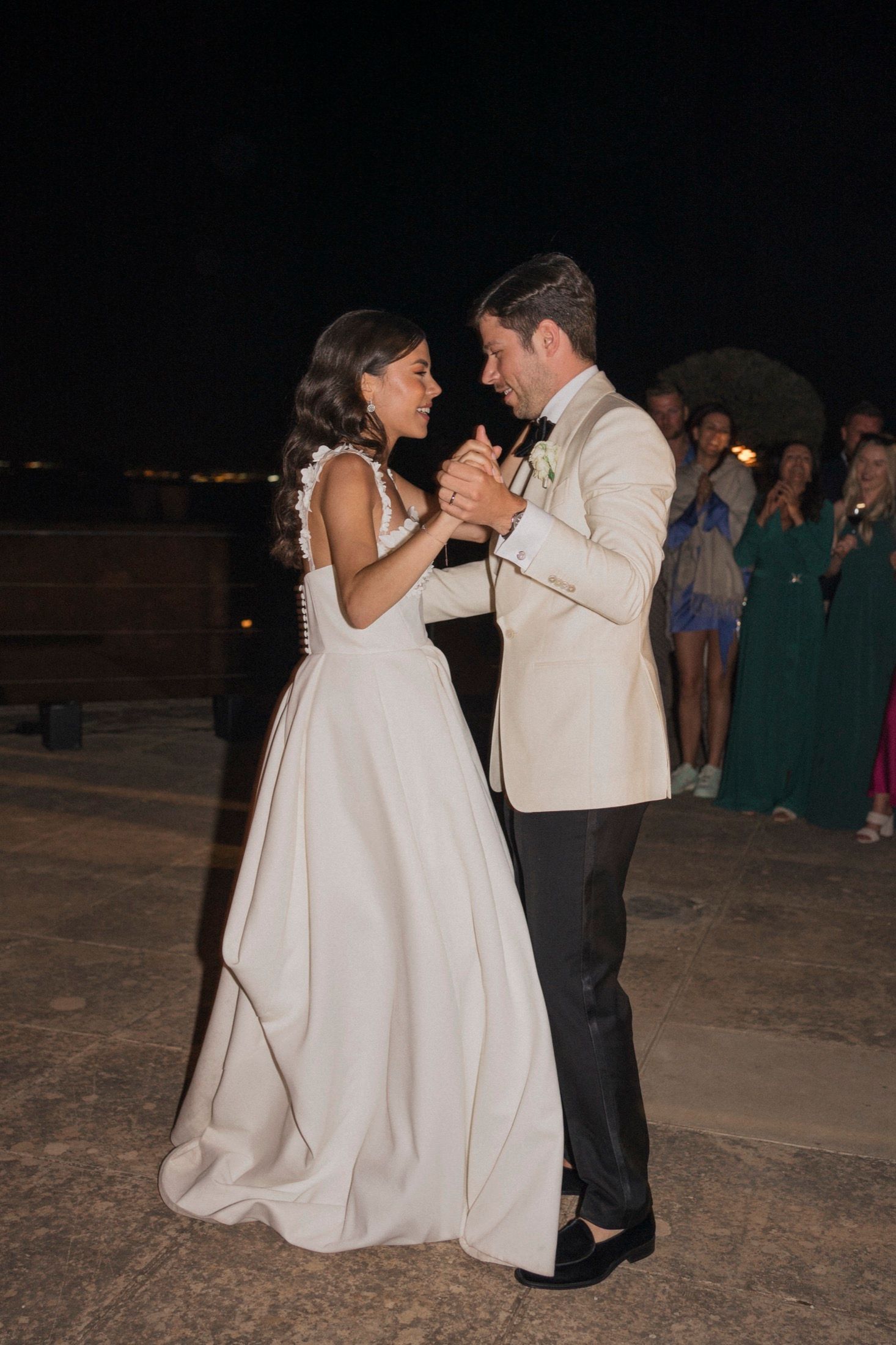 Bride and groom share their first dance outdoors at night, she in a white gown, he in a cream tuxedo jacket.