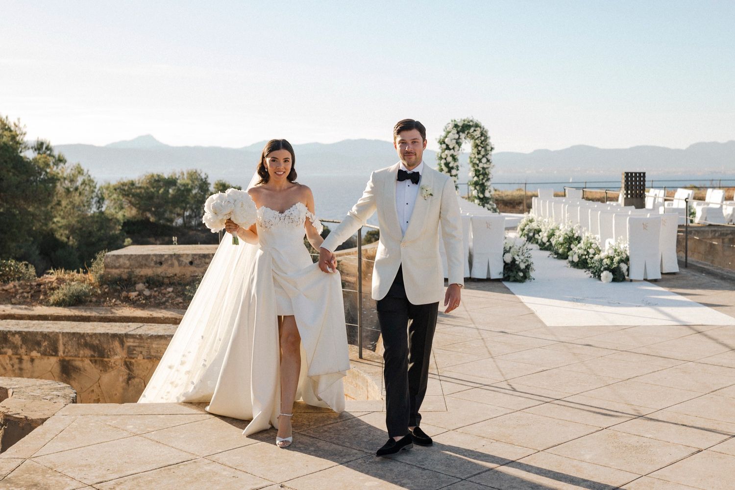 Bride and groom walking hand in hand at an outdoor wedding venue with mountain views and floral arch.