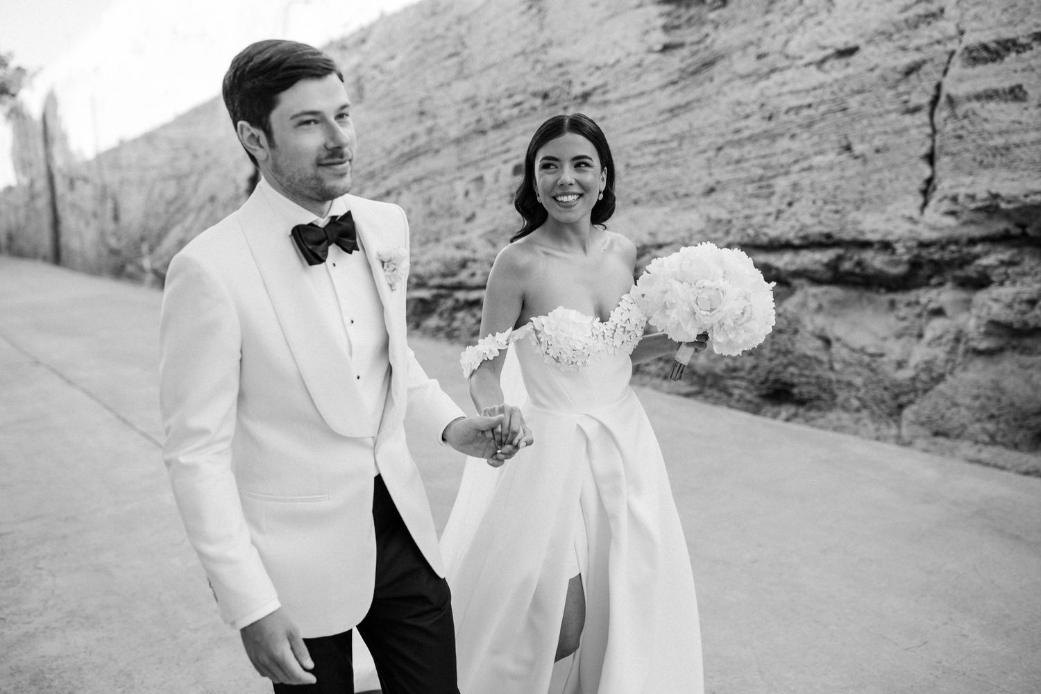 Black and white photo of a smiling bride holding white bouquet and groom in tuxedo walking outdoors near rock wall.