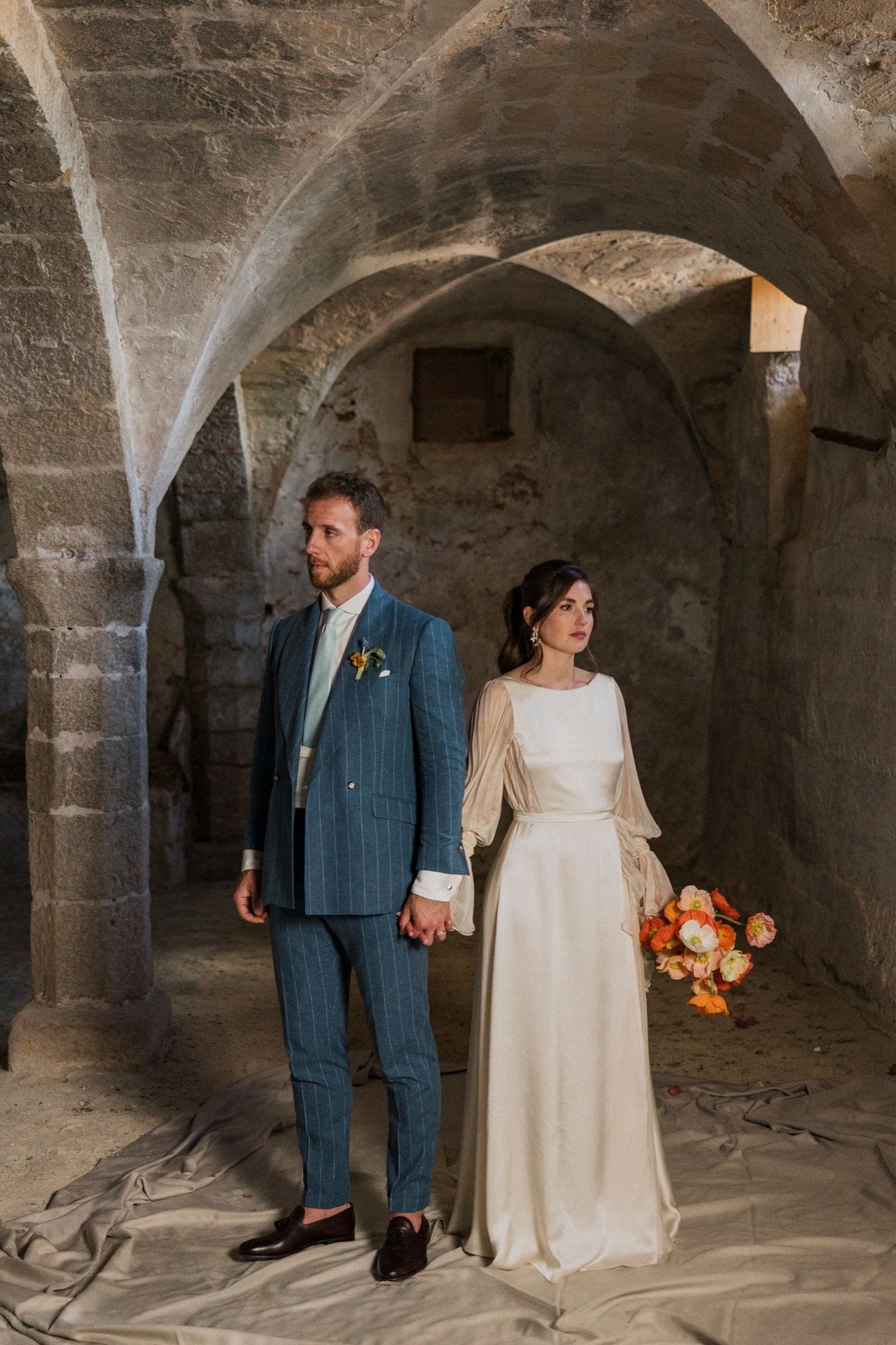 A couple in wedding attire stands in a stone archway tunnel holding hands and carrying an orange floral bouquet.