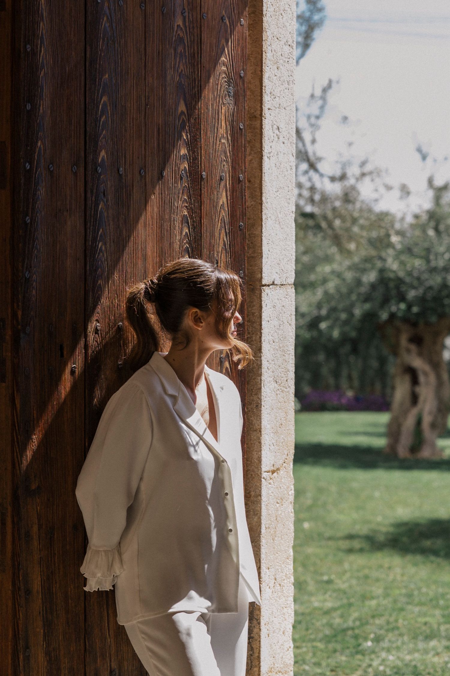 Person in all-white outfit leans against rustic wooden door frame in garden setting with sunlit natural backdrop.