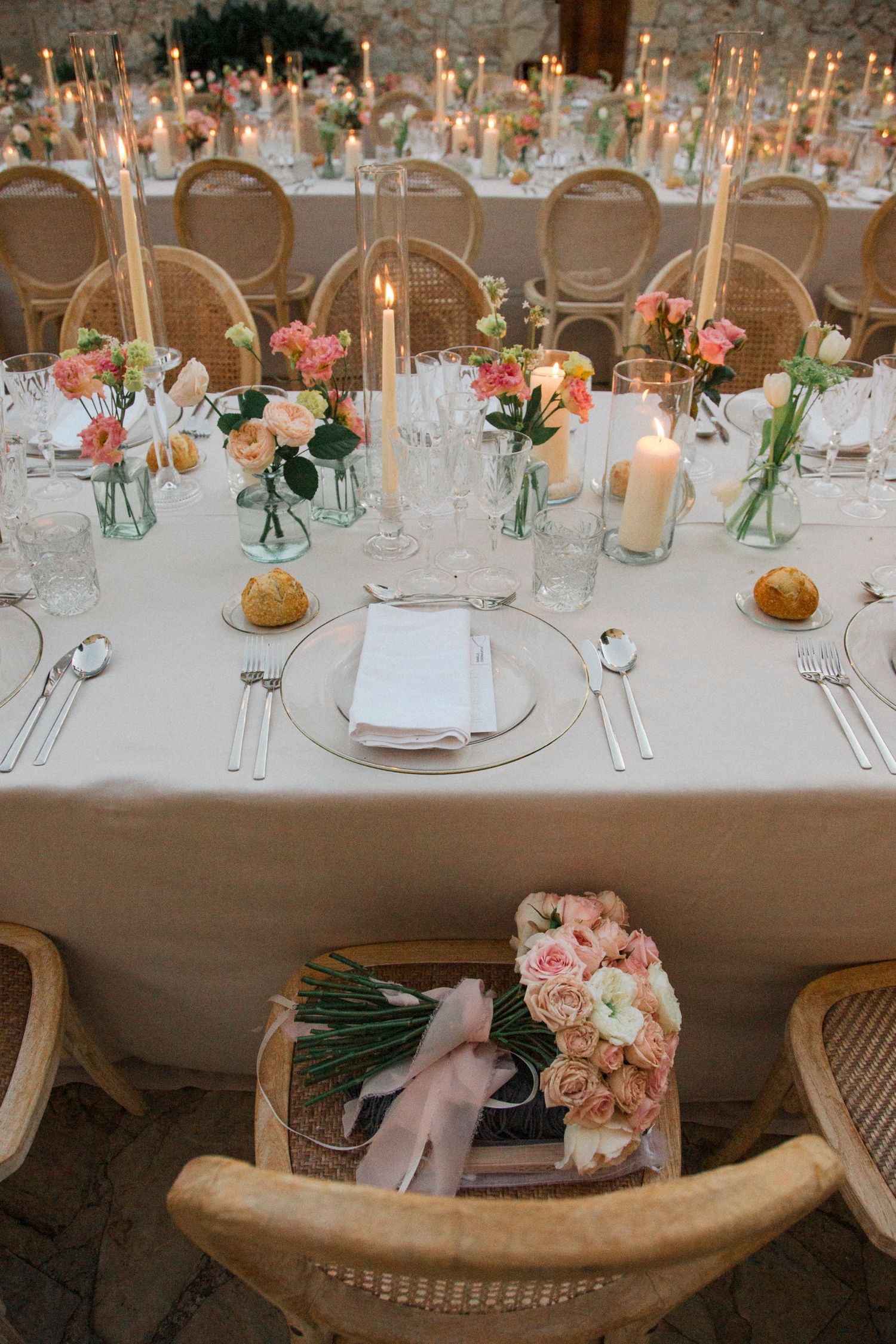 Elegant wedding reception table settings with pink floral centerpieces, glassware, and woven chairs in a banquet hall.