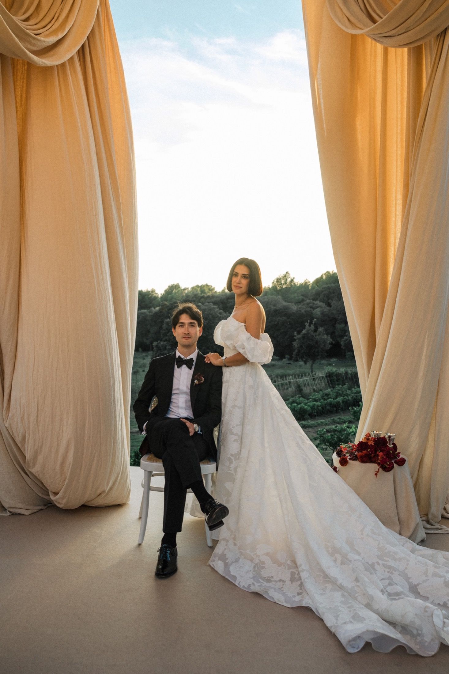 A wedding couple poses against elegant cream-colored curtains in a romantic outdoor setting at sunset.