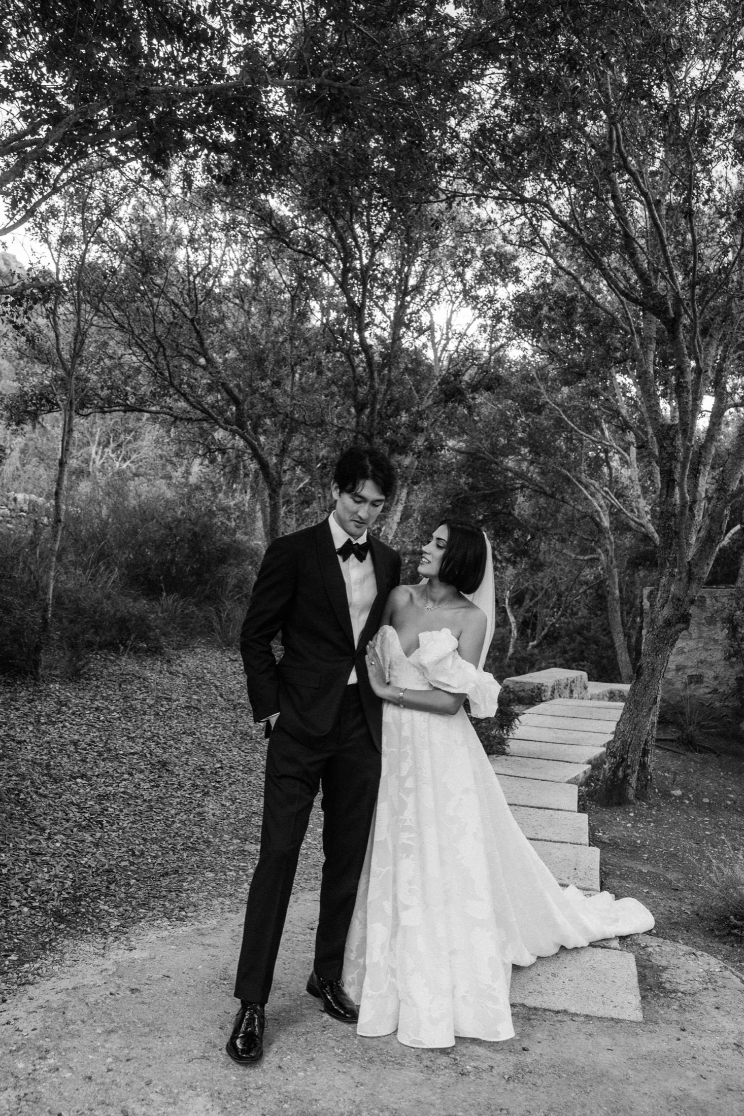 A couple in formal wedding attire stand together on stone steps surrounded by trees in a black and white photo.