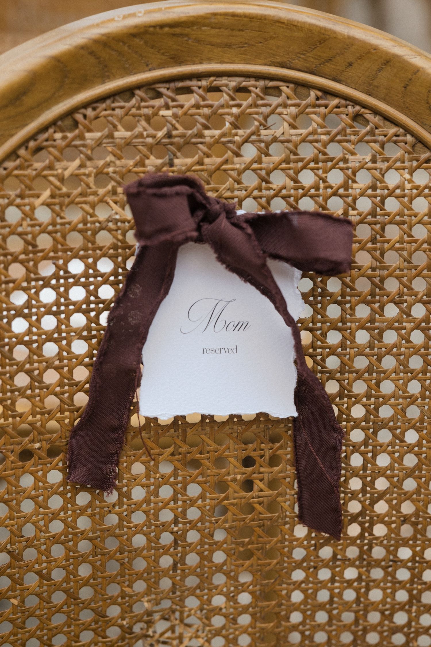 A white menu card with elegant script text tied with a brown satin bow on a vintage cane-backed chair.