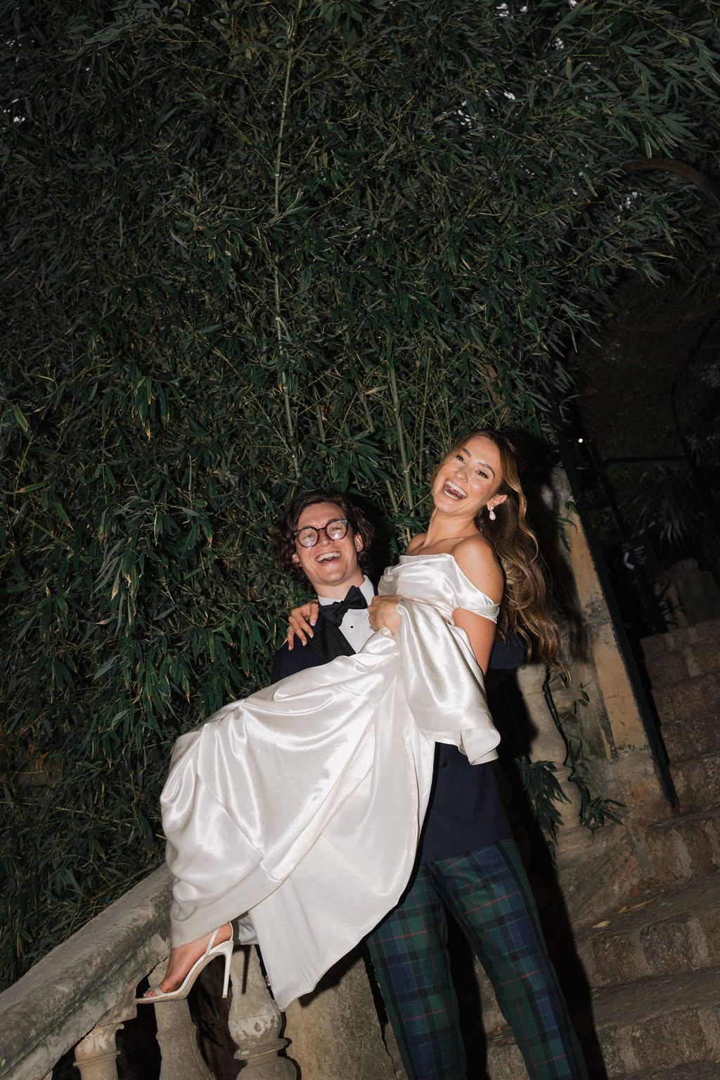 Playful wedding couple posing on stairs against bamboo backdrop at night.