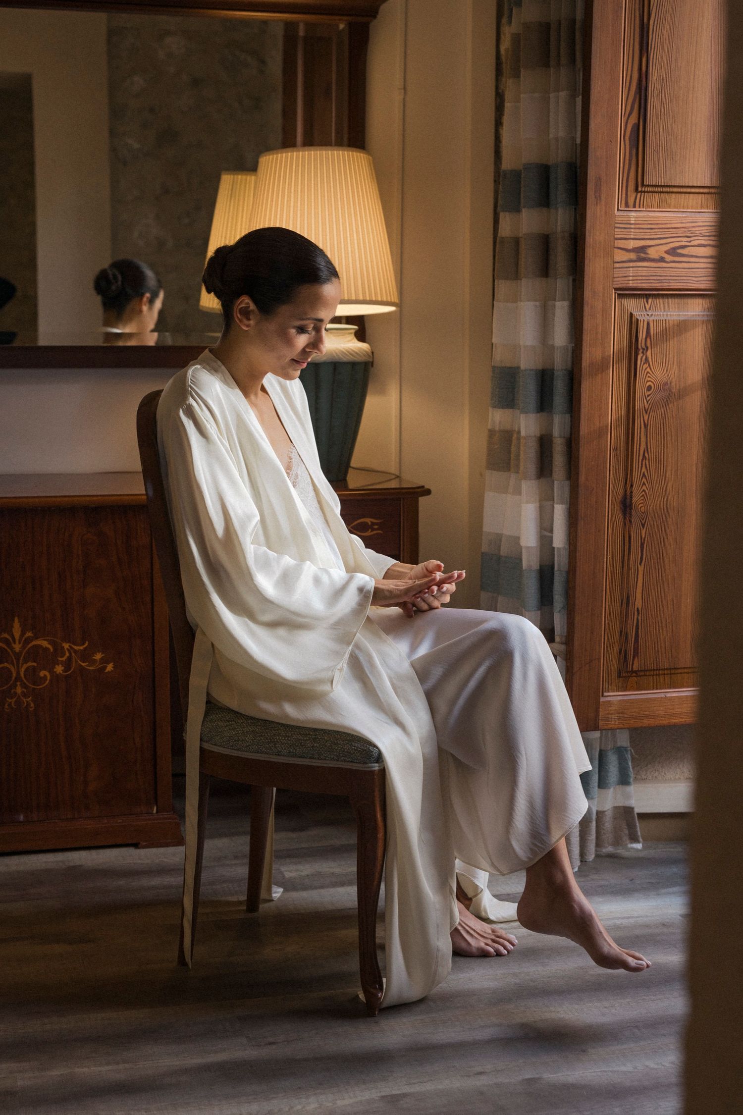 Person in white dress sits thoughtfully on chair in sunlit room with wooden details.