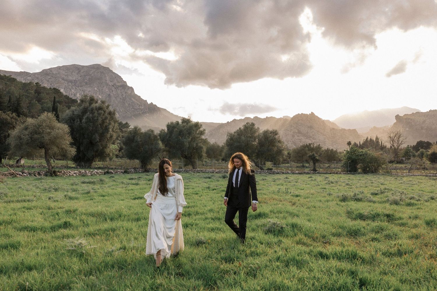 A couple walks through a lush green meadow with dramatic mountains and cloudy skies in the background.