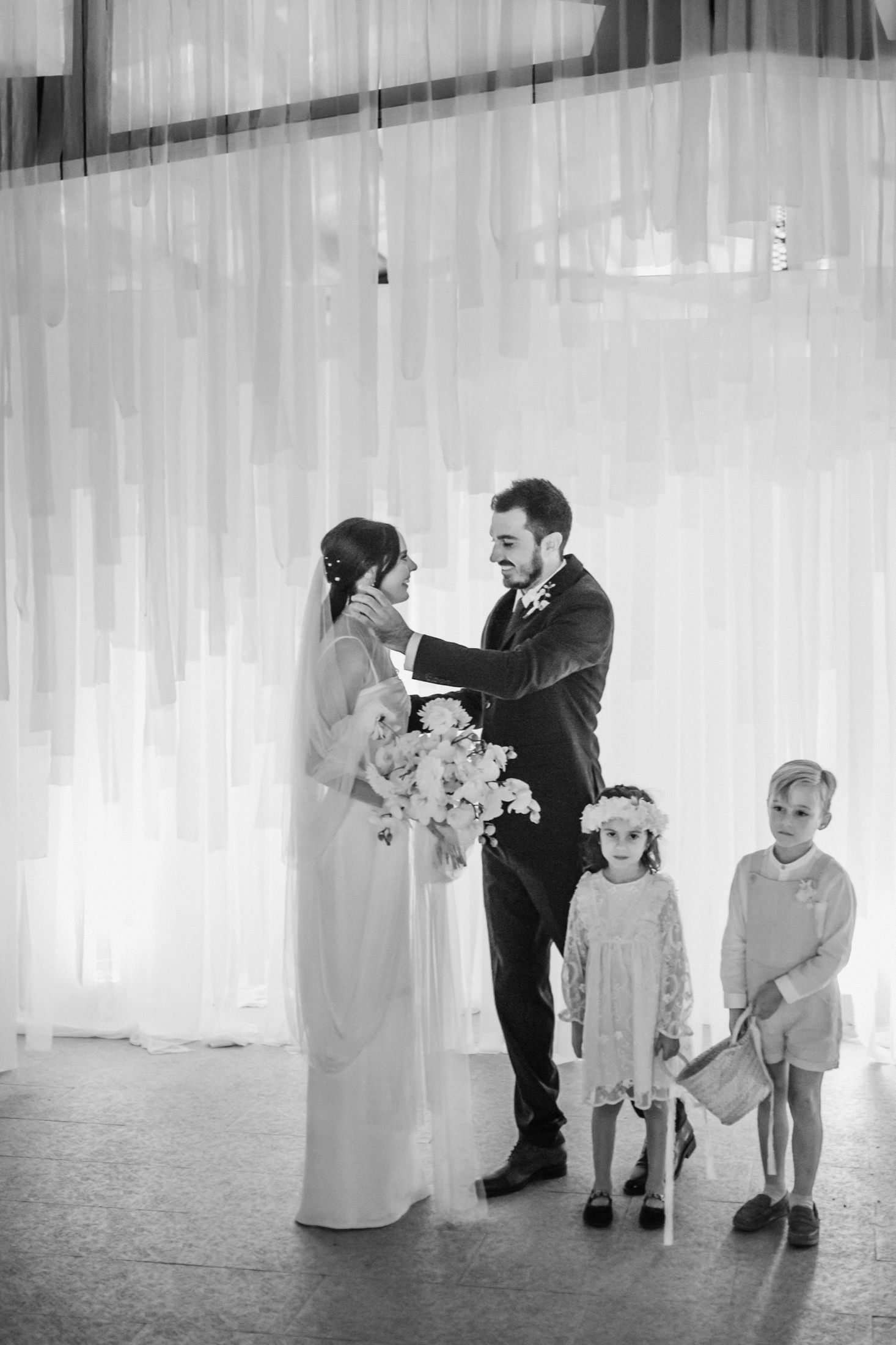 Black and white wedding reception scene with children playing near sheer white curtains.
