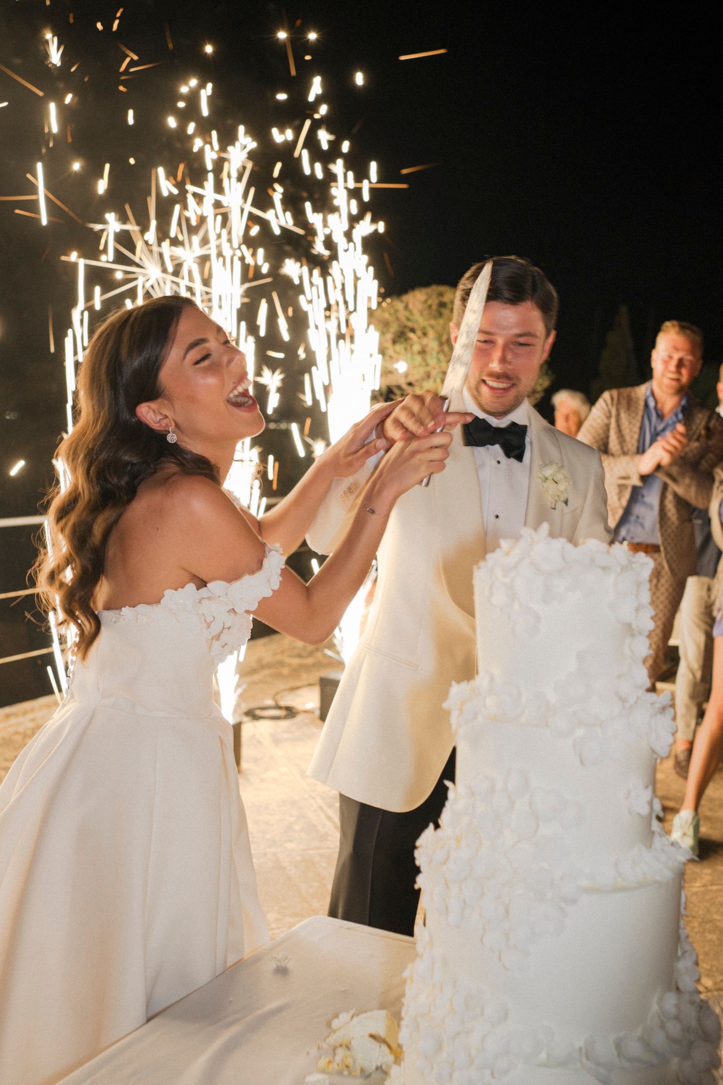 Bride and groom cutting white wedding cake with sparklers at night outdoor reception.