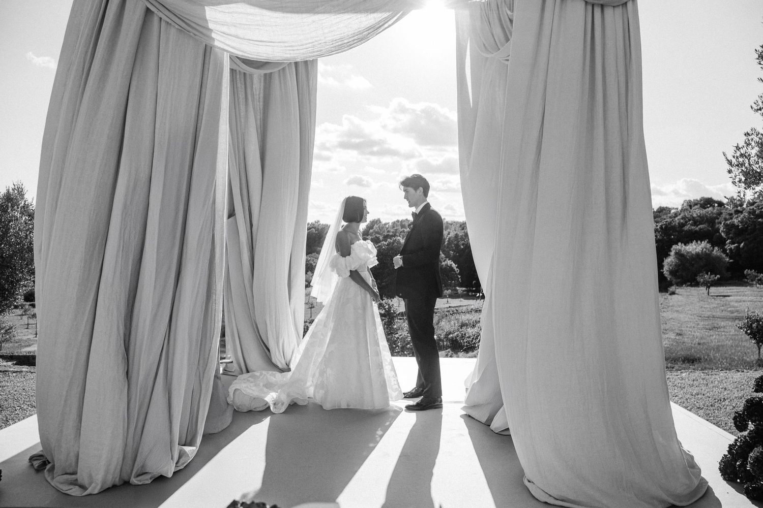 Black and white wedding ceremony photo showing couple at altar under flowing white fabric drapes outdoors.