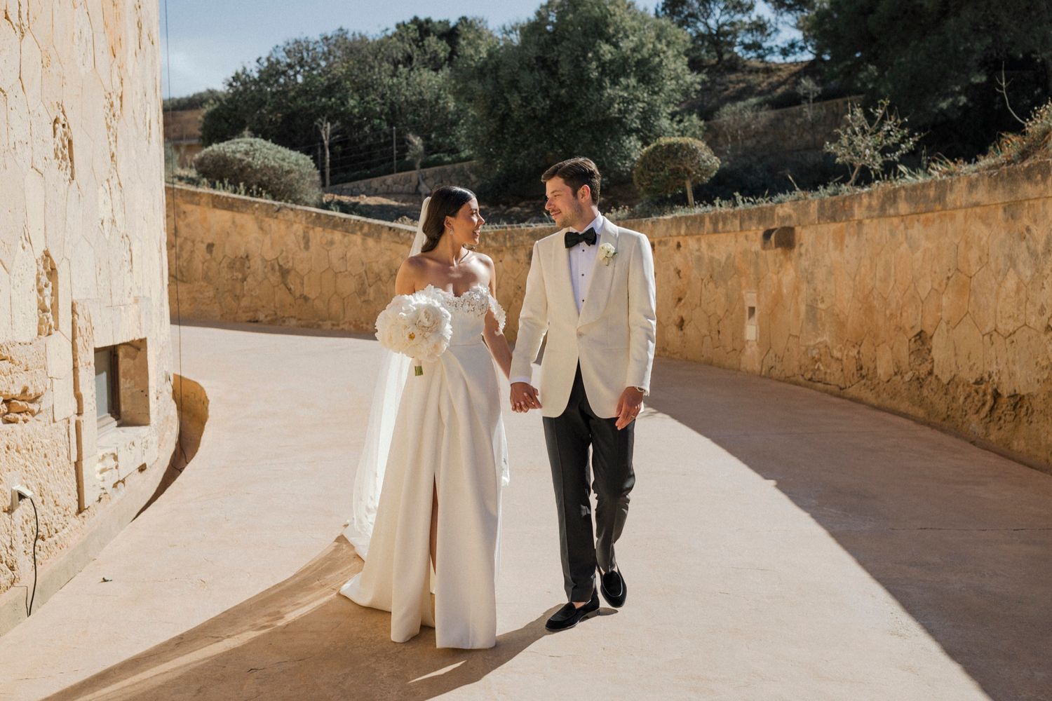 Bride and groom walk hand-in-hand along a sunlit stone pathway, bride in elegant off-shoulder gown, groom in white tuxedo.