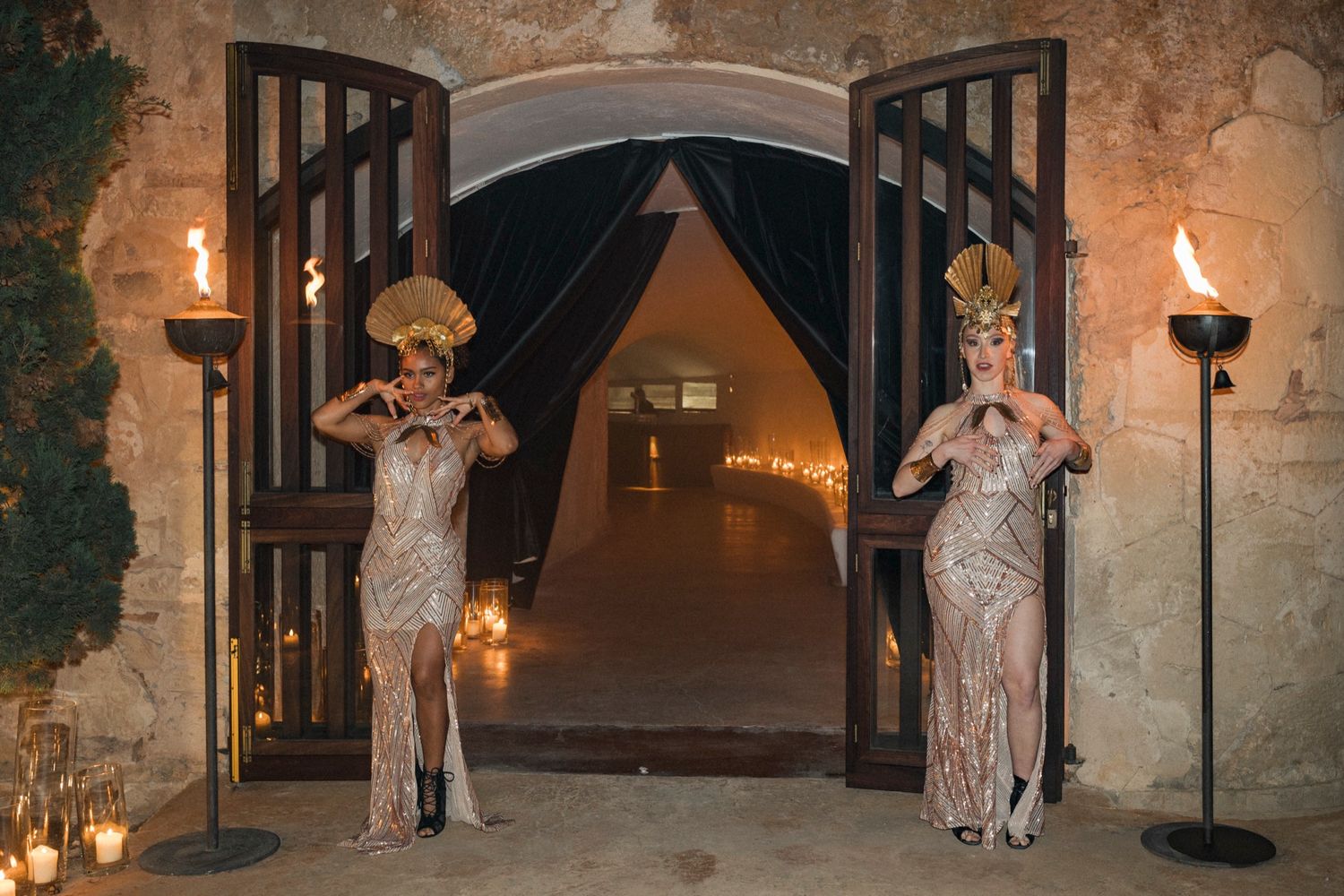 Two glamorous performers in gold sequin gowns and Egyptian headdresses pose at torch-lit stone archway entrance.