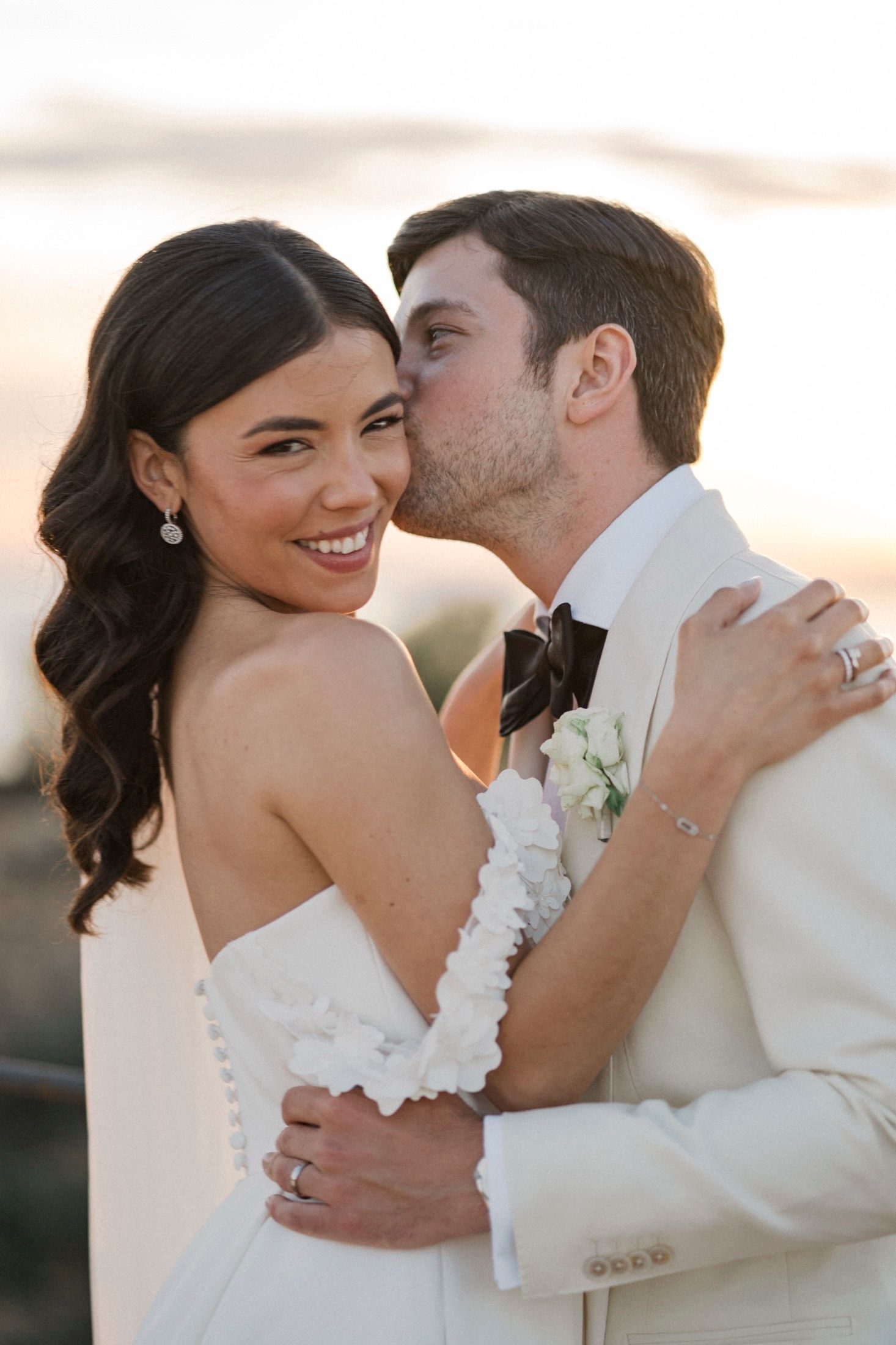 Groom in white tuxedo kissing bride on cheek as she smiles at camera during golden hour wedding portrait.