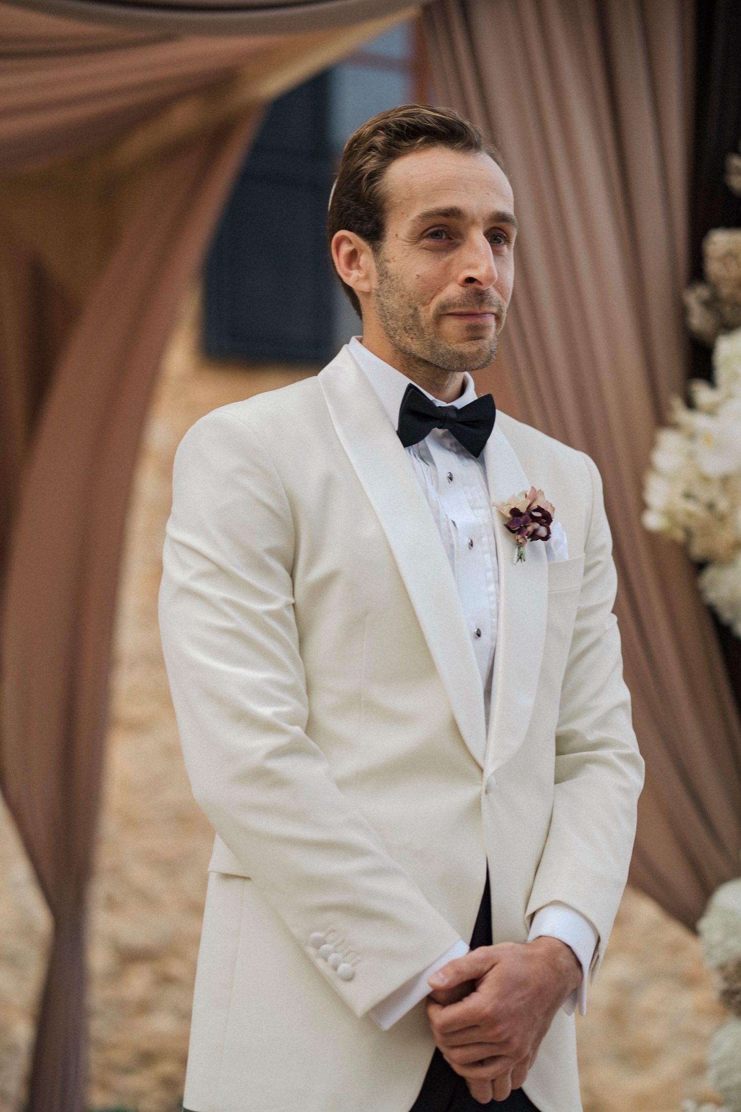 A groom in a classic white tuxedo jacket and black bow tie smiles during a wedding ceremony.