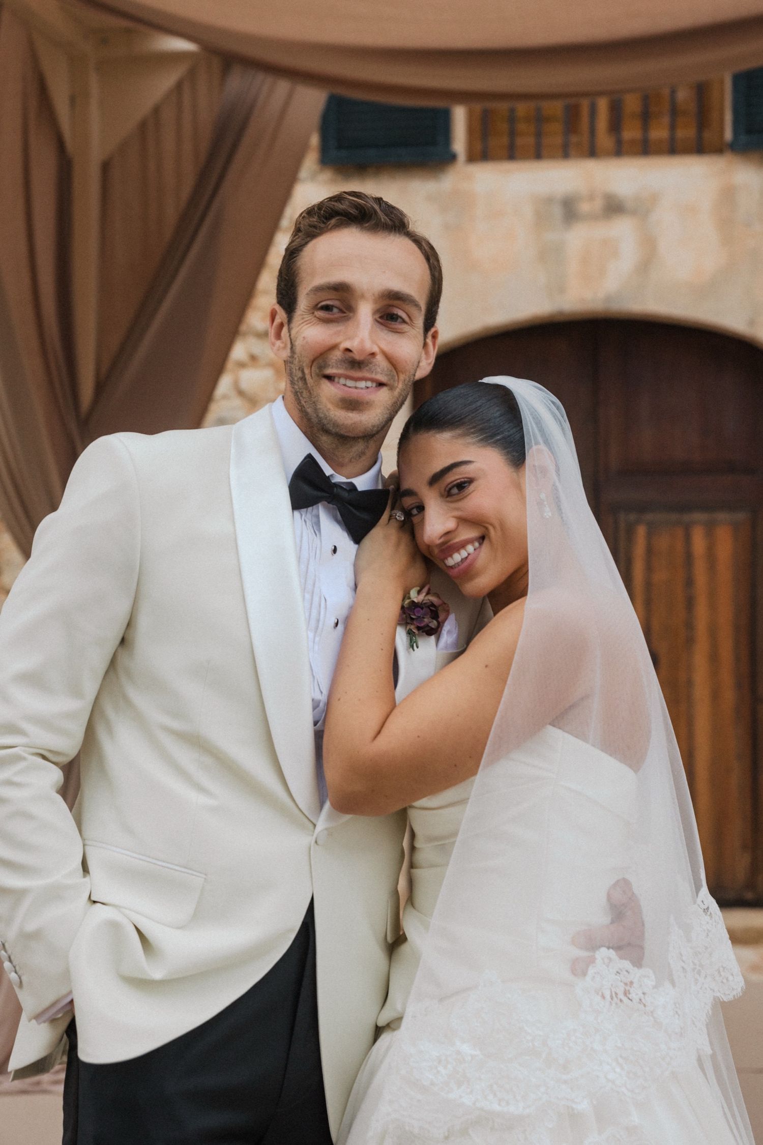 A happy wedding couple embraces and smiles together in formal white attire and a veil.