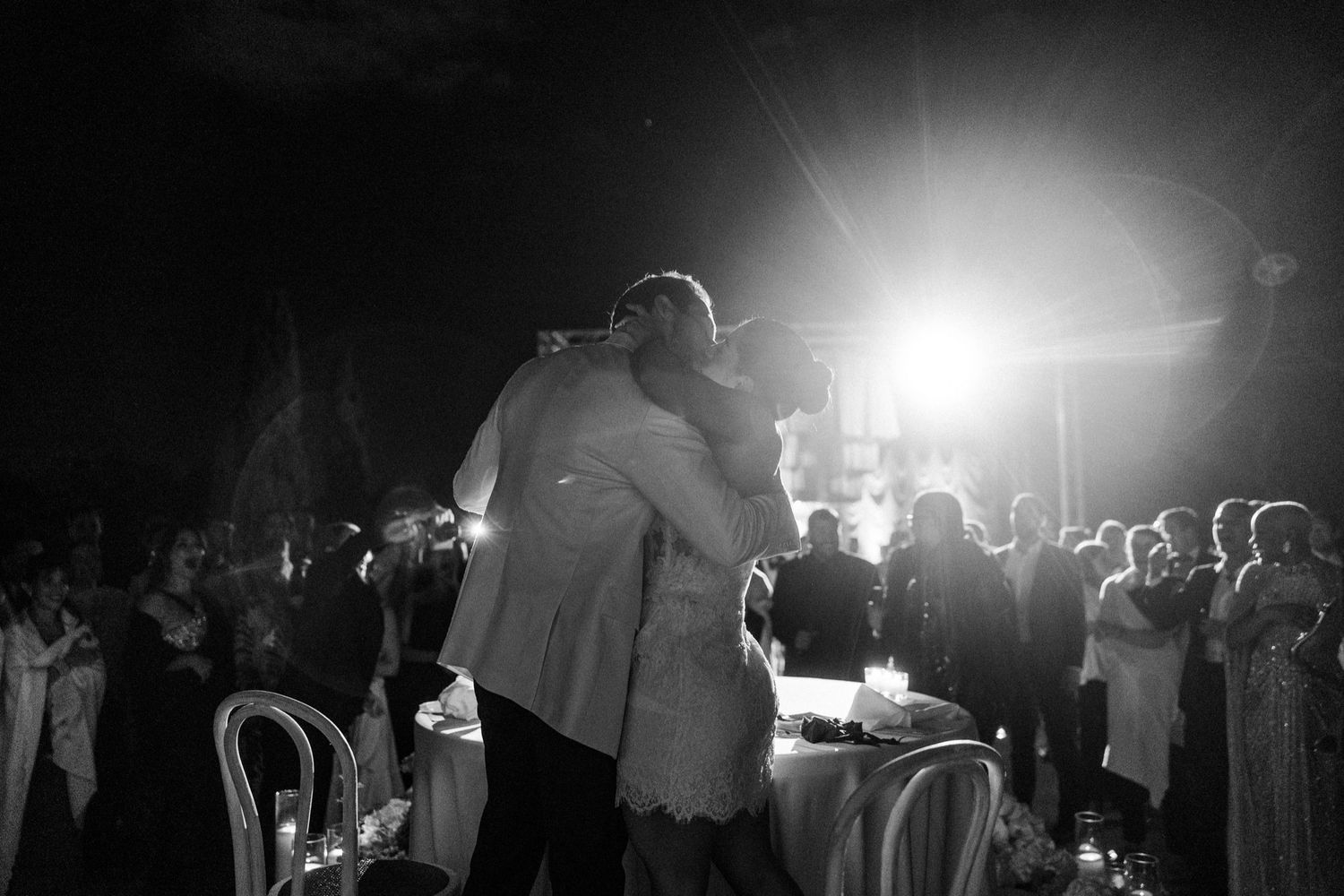 A black and white photo of a nighttime wedding ceremony with bright backlighting and silhouetted figures gathered around.