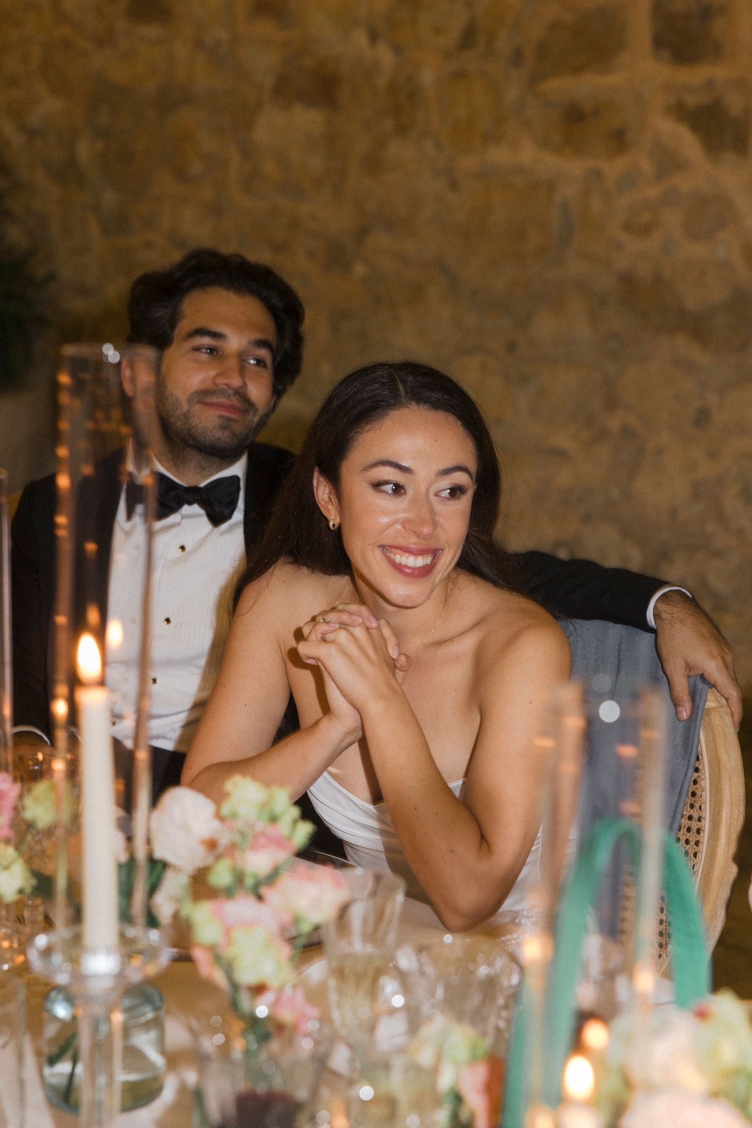 Wedding reception guests share laughter and smiles at an elegant stone-walled venue with floral centerpieces and candlelight.