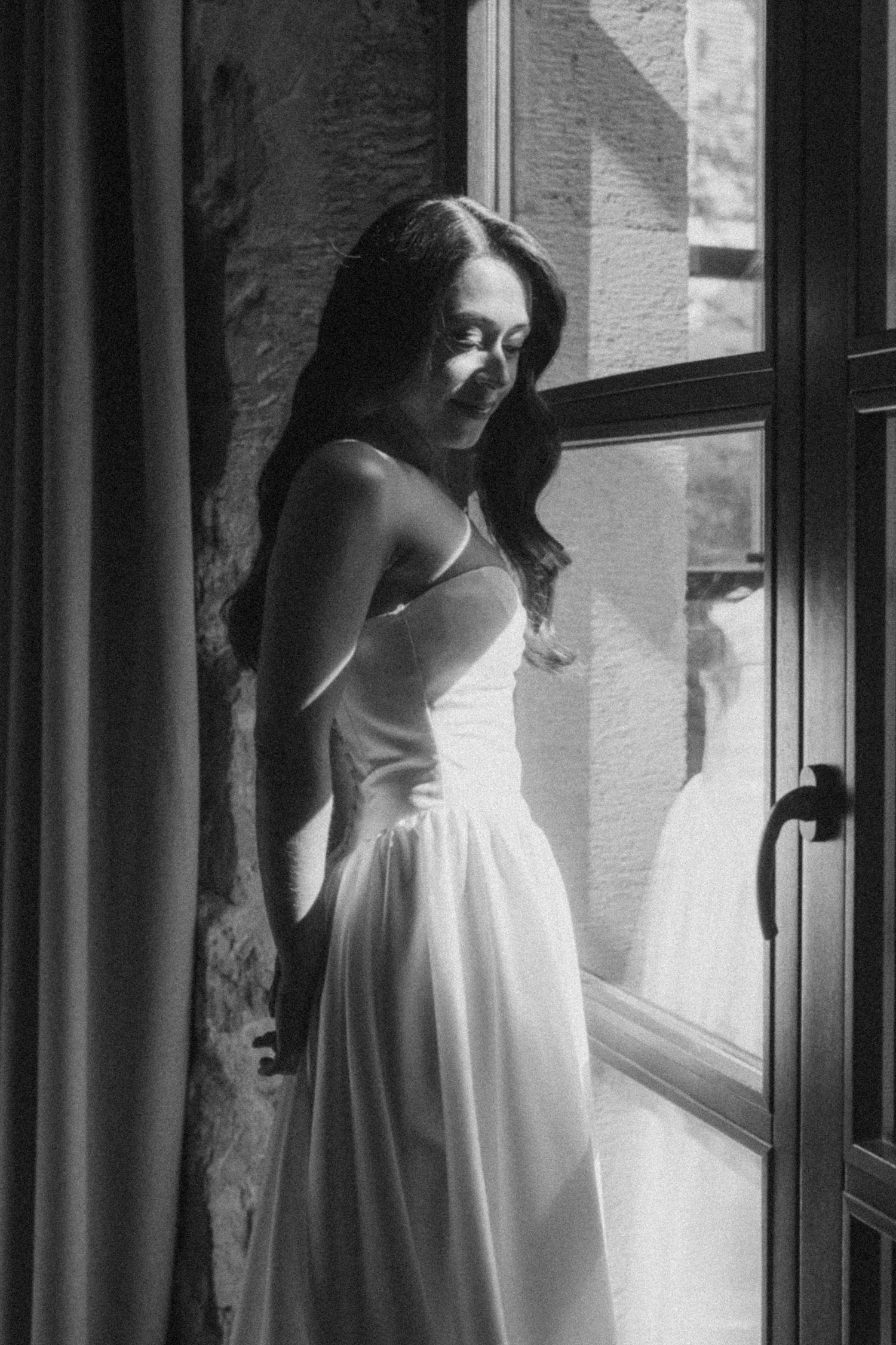 Black and white artistic portrait of a woman in a white dress standing by a window, creating multiple reflections.