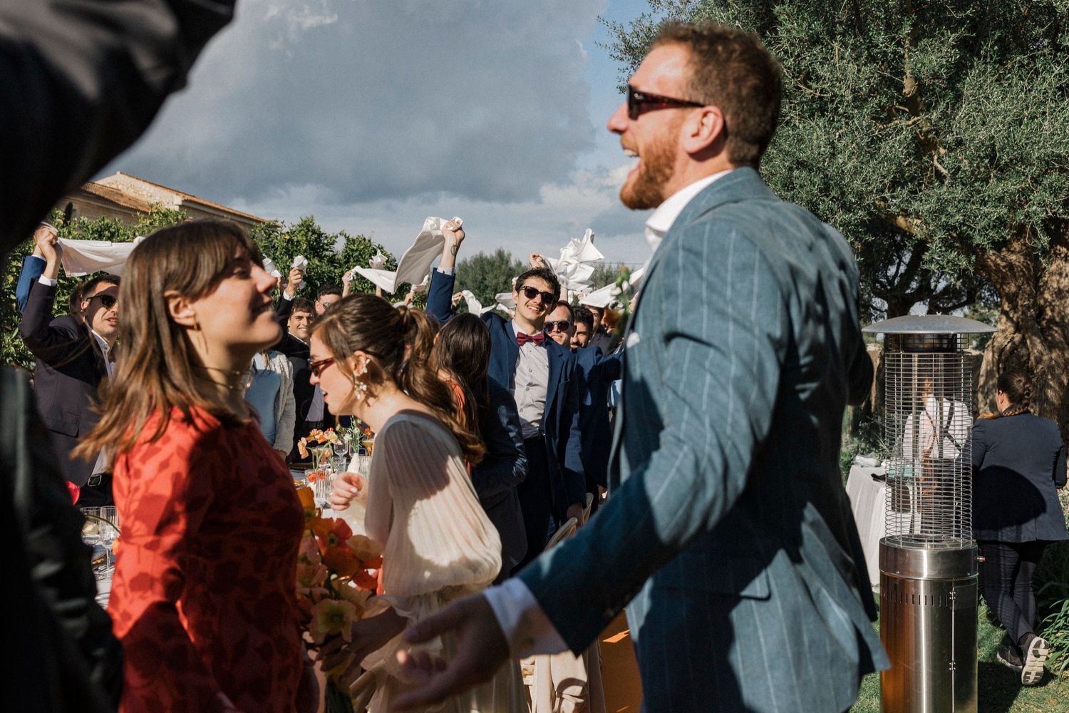 A group of people gather outdoors for a social celebration in a natural setting with trees and sunlight.