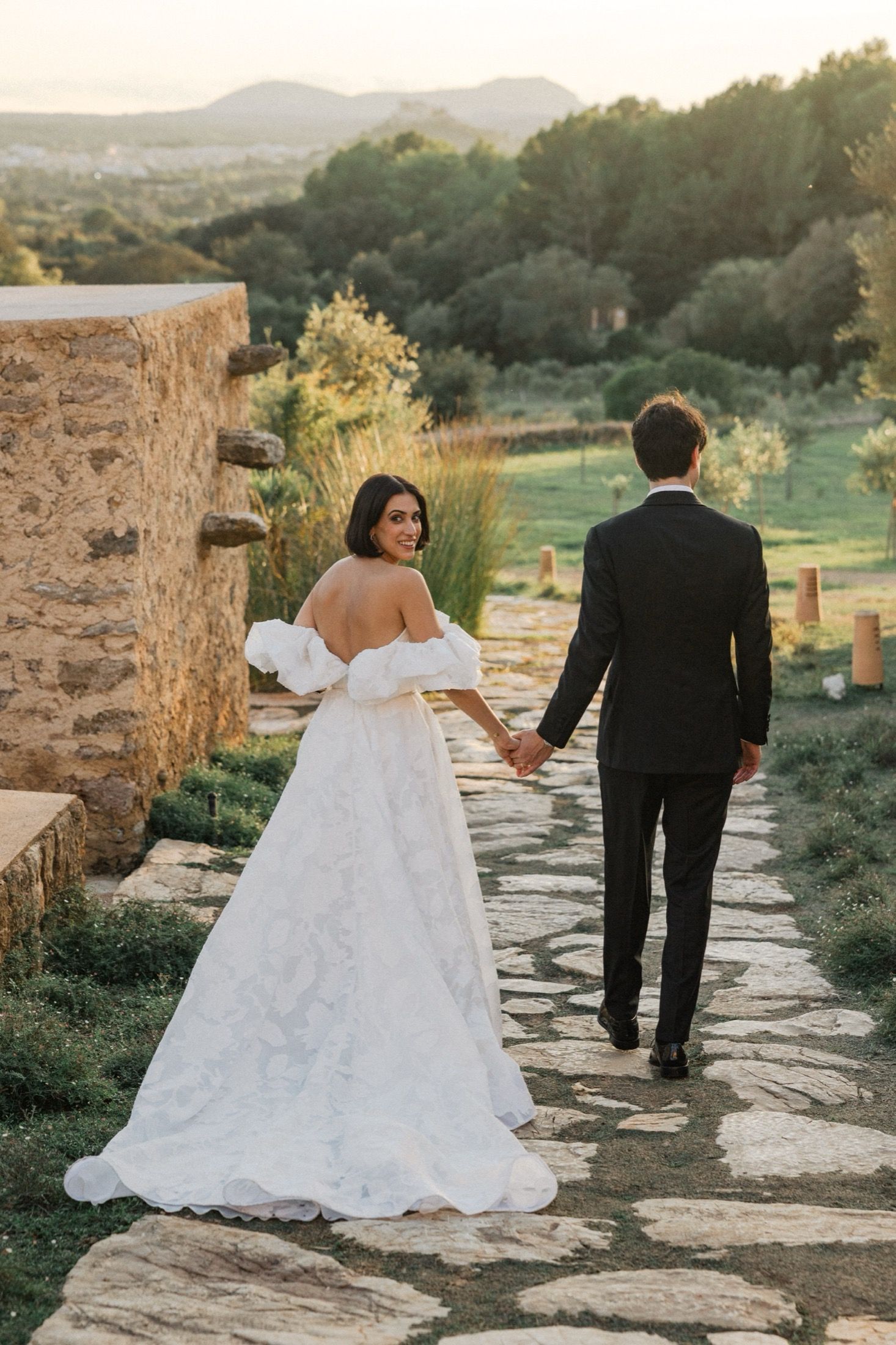 A couple in wedding attire walks along a stone path at sunset with mountains and greenery in the background.