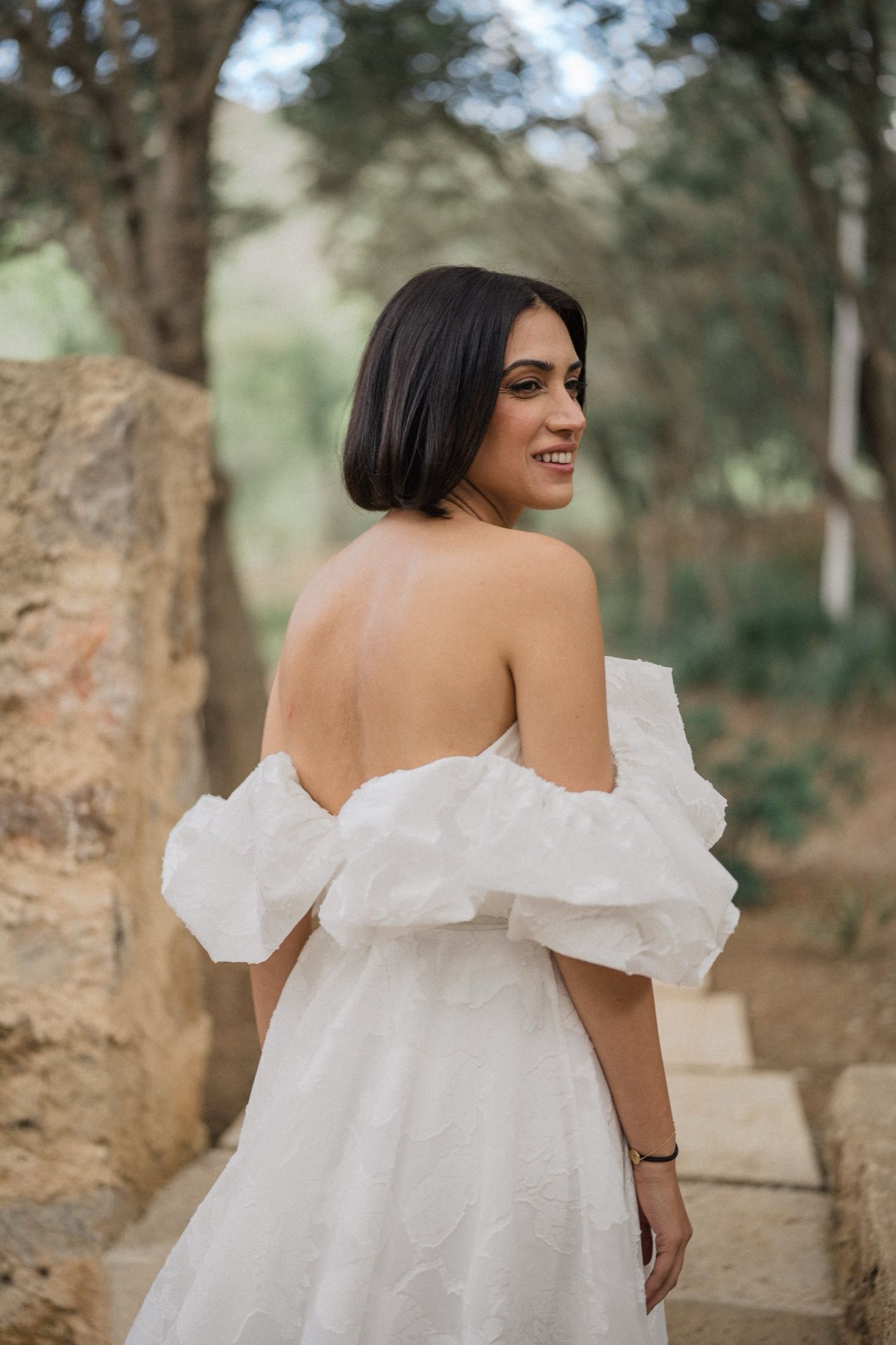 Woman in a flowing white off-shoulder dress poses outdoors in a natural setting with trees in the background.