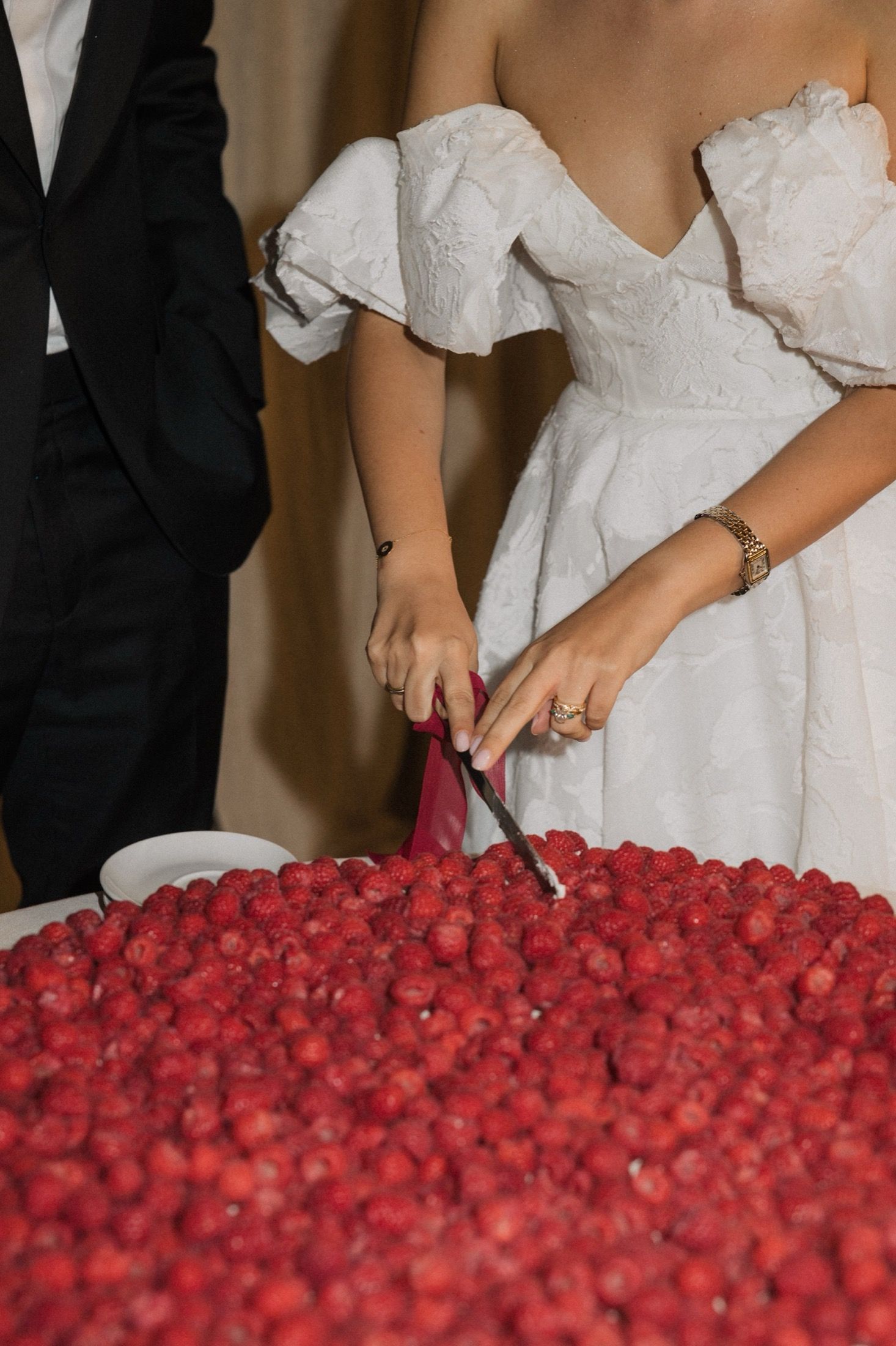 A person in a white wedding dress cutting into a large cake decorated with vibrant red raspberries.