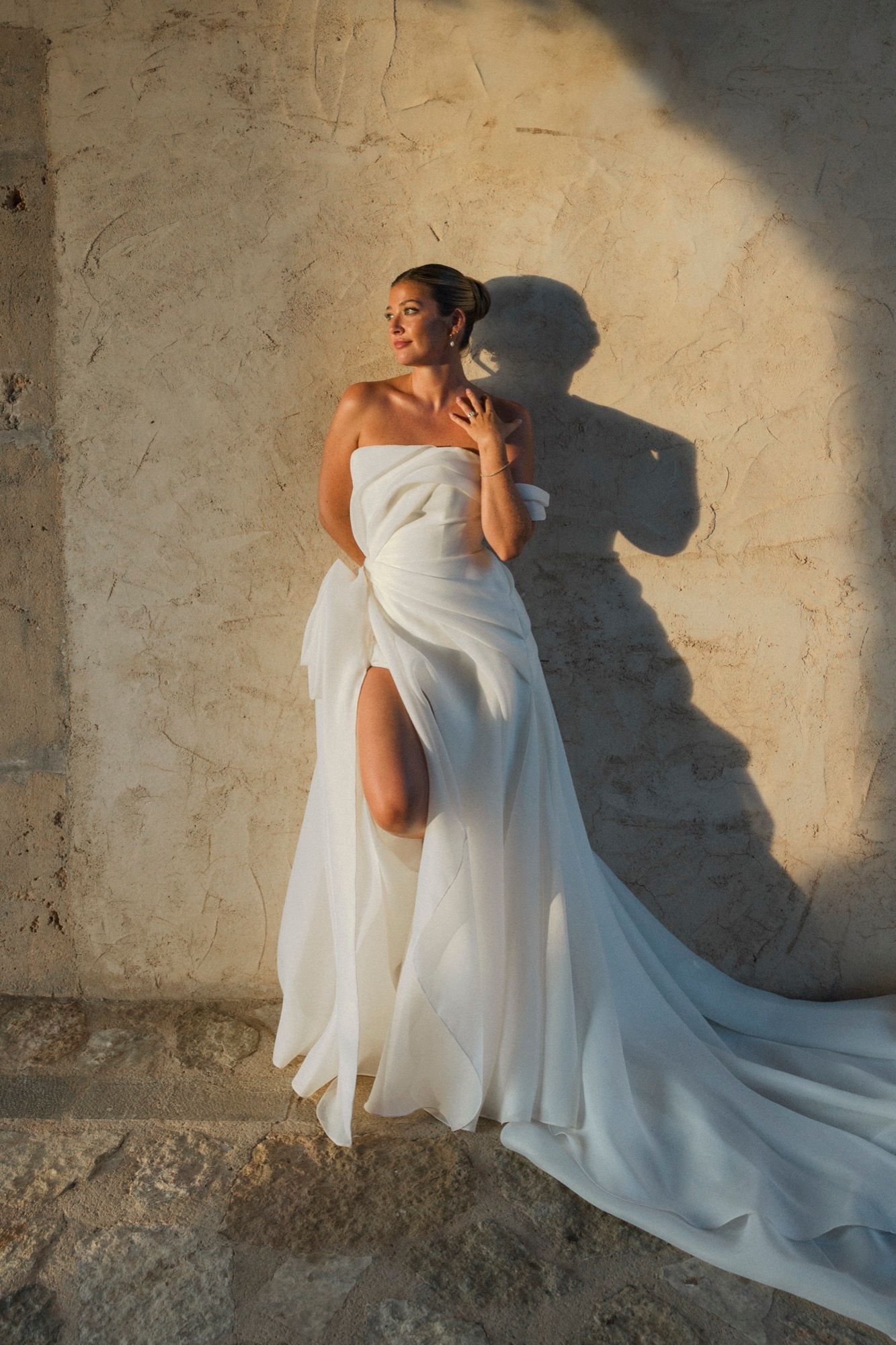 A bride in an elegant white gown poses dramatically against a sunlit stone wall.