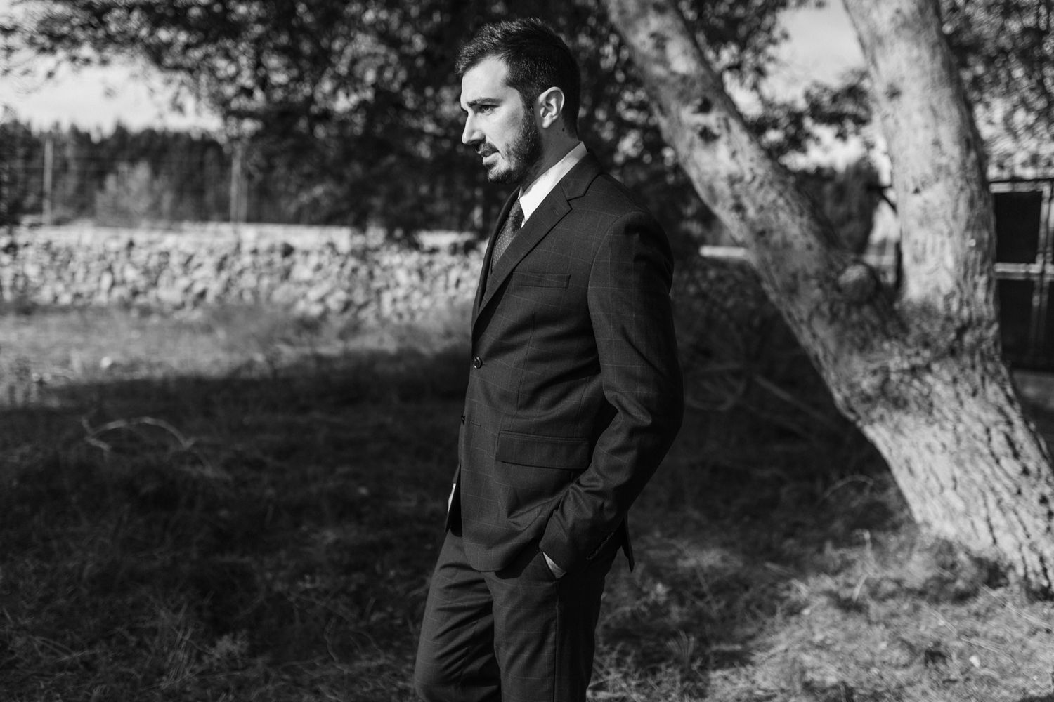 Black and white photo of someone in a dark suit standing outdoors near trees with hands in pockets.