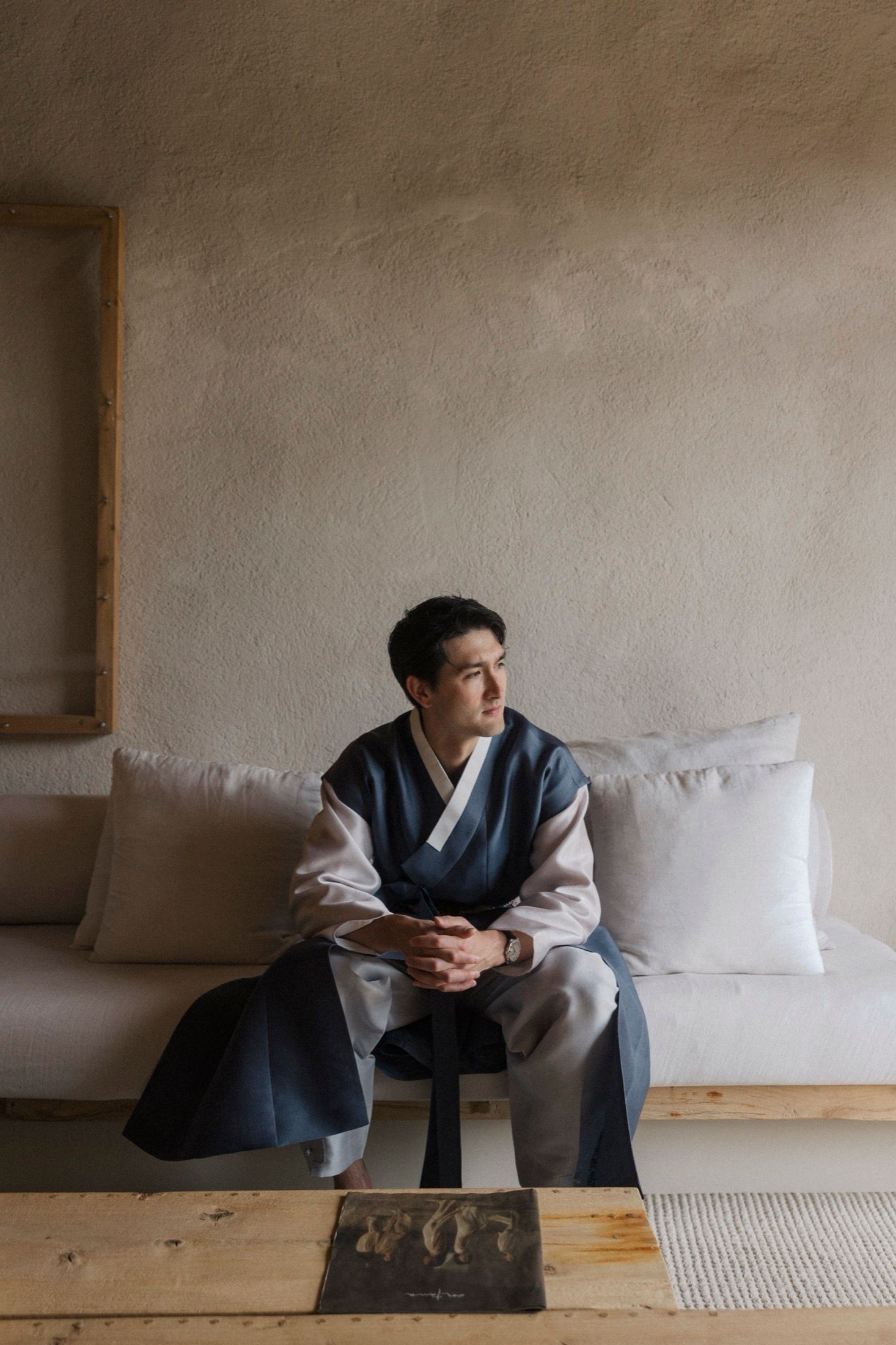A person in traditional Korean hanbok attire sits contemplatively on a wooden floor against a plain wall.