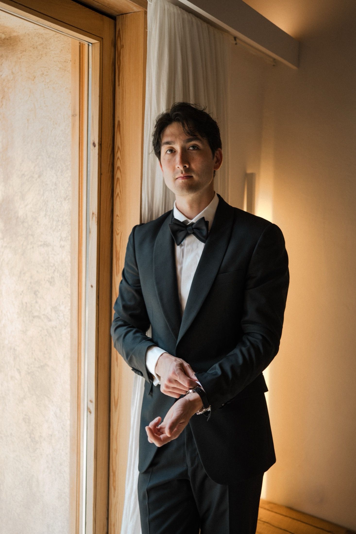 A person in a black suit stands in a warmly lit hallway against a beige wall.