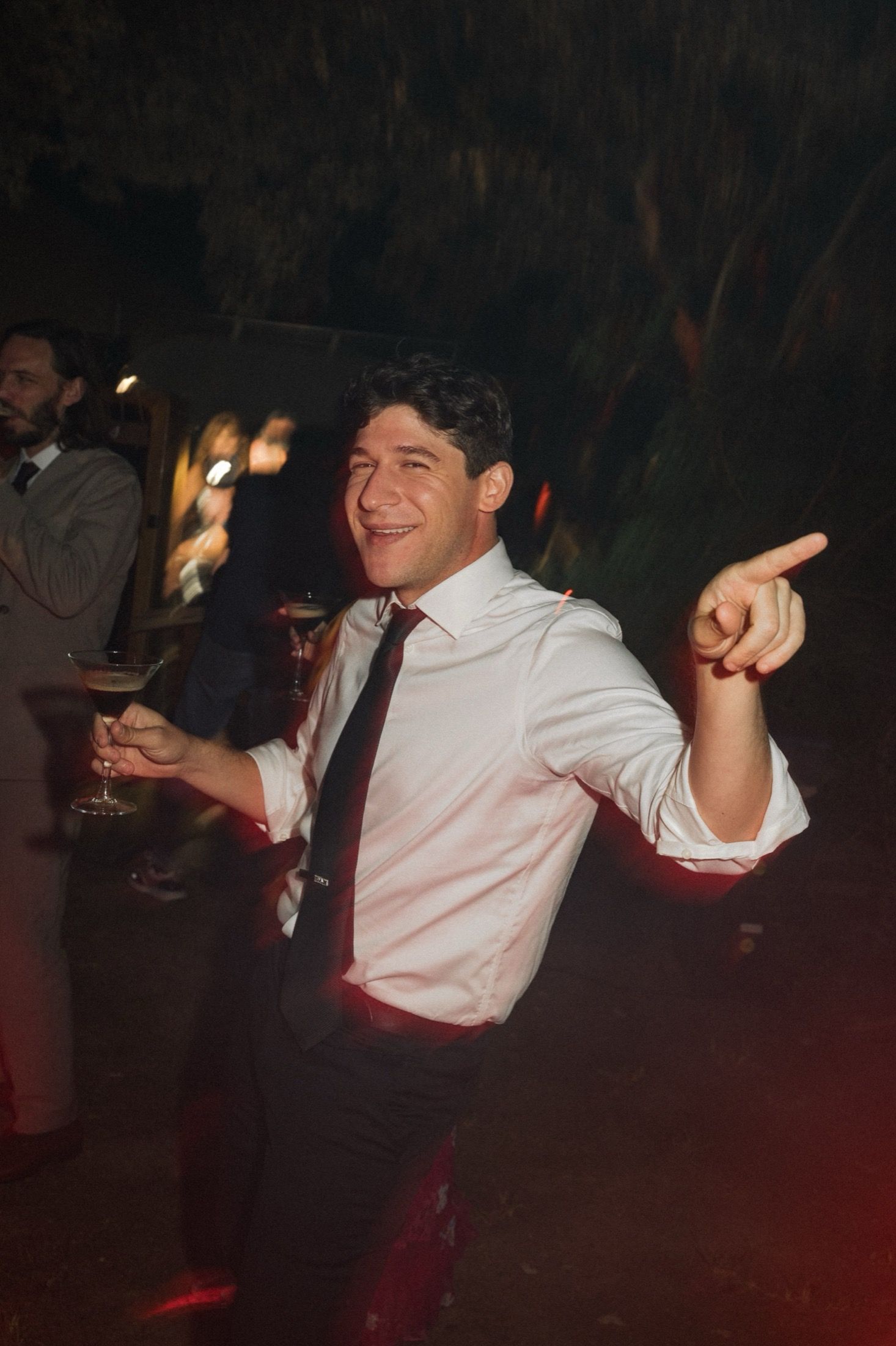 A person in a white dress shirt and tie dances joyfully at a nighttime party while holding a drink.