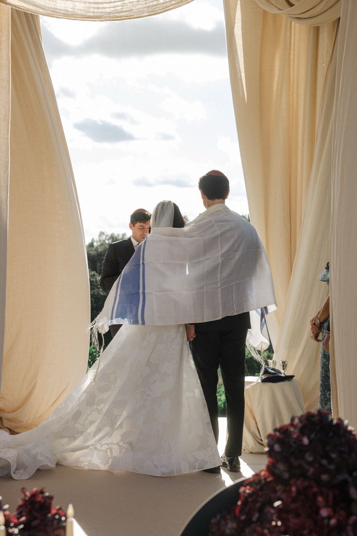A couple stands under draped fabric at an outdoor wedding ceremony with burgundy floral decorations visible.