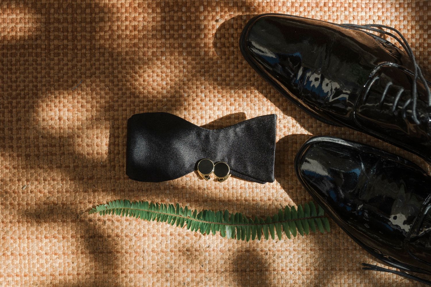 A black bow tie and shiny dress shoes arranged on a textured brown surface with dappled lighting.