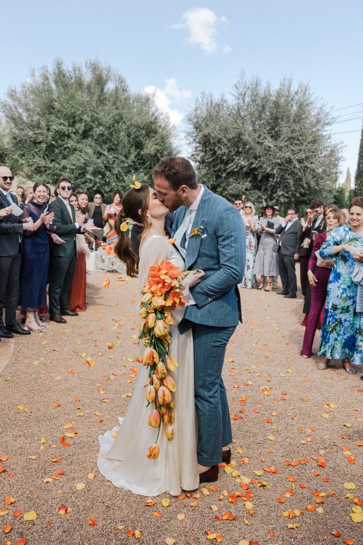 A couple shares a romantic kiss at an outdoor wedding ceremony while guests look on and orange petals scatter around them.