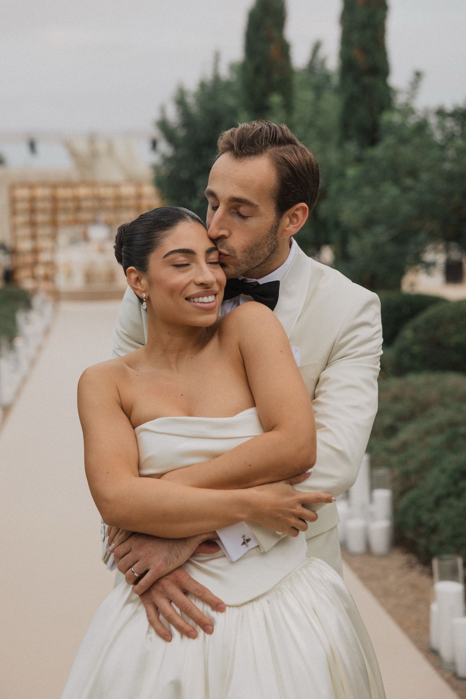 A romantic wedding couple embraces outdoors, with the bride in a white gown and groom in cream suit.