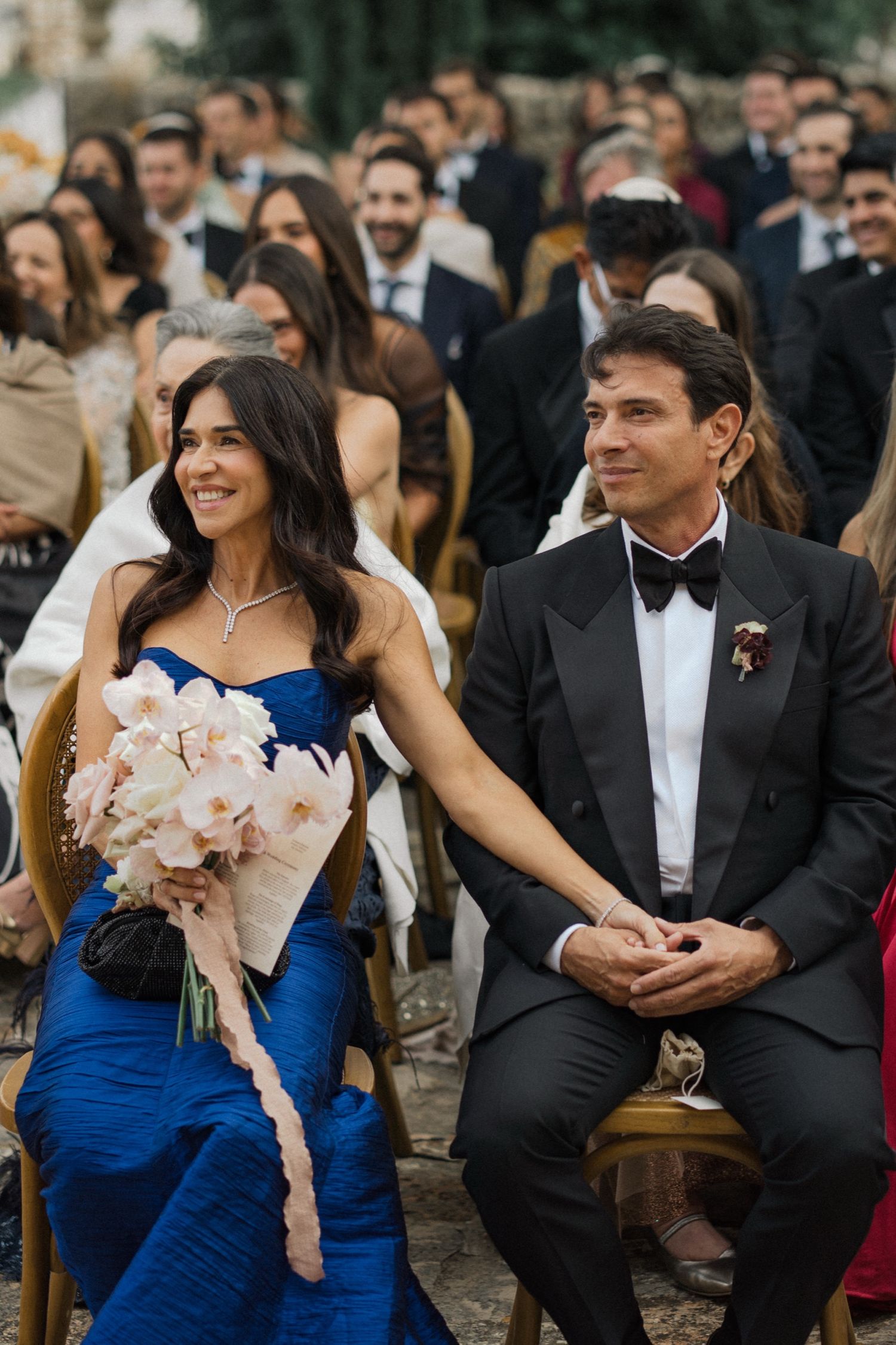 Wedding guests in formal attire sit outdoors as bridesmaids in blue dresses and groomsmen in tuxedos socialize at reception.