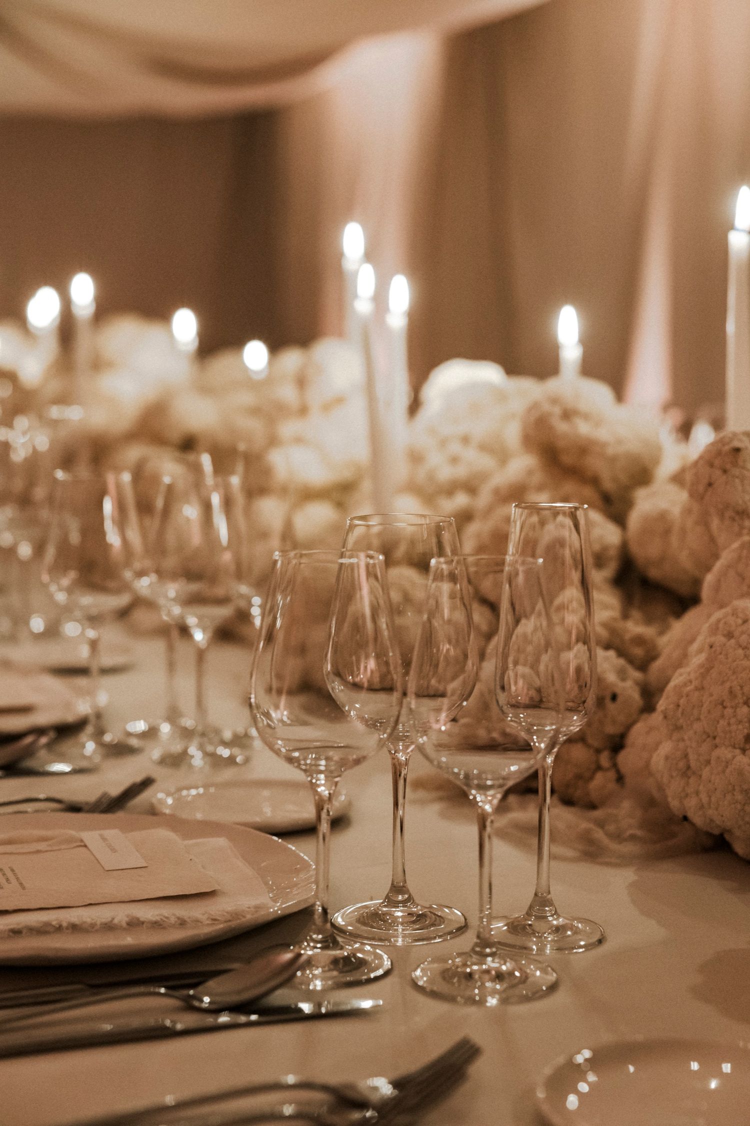 Close-up of formal place settings with wine glasses and floral centerpieces in warm lighting.