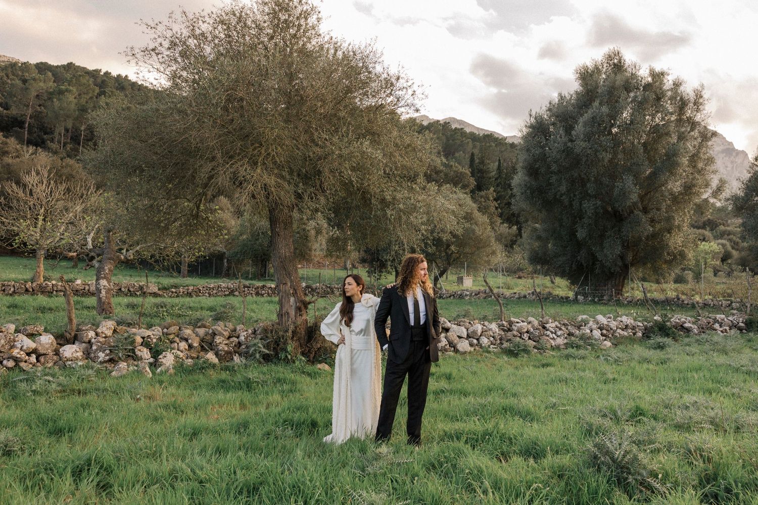 A wedding couple poses in a rustic field with stone walls and mountains creating a scenic backdrop.