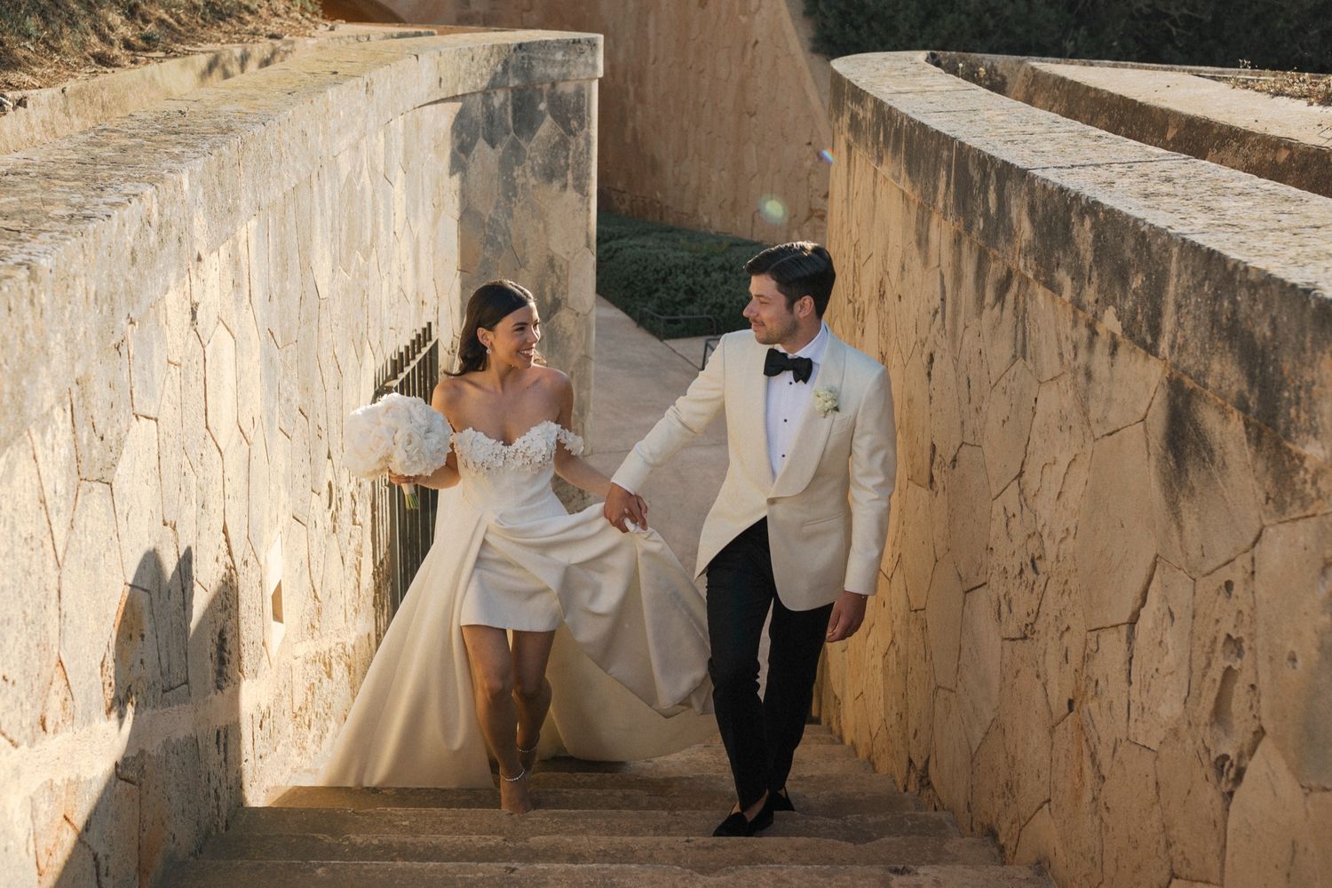 Bride in white mini dress and groom in ivory tuxedo hold hands walking up stone steps at golden hour.