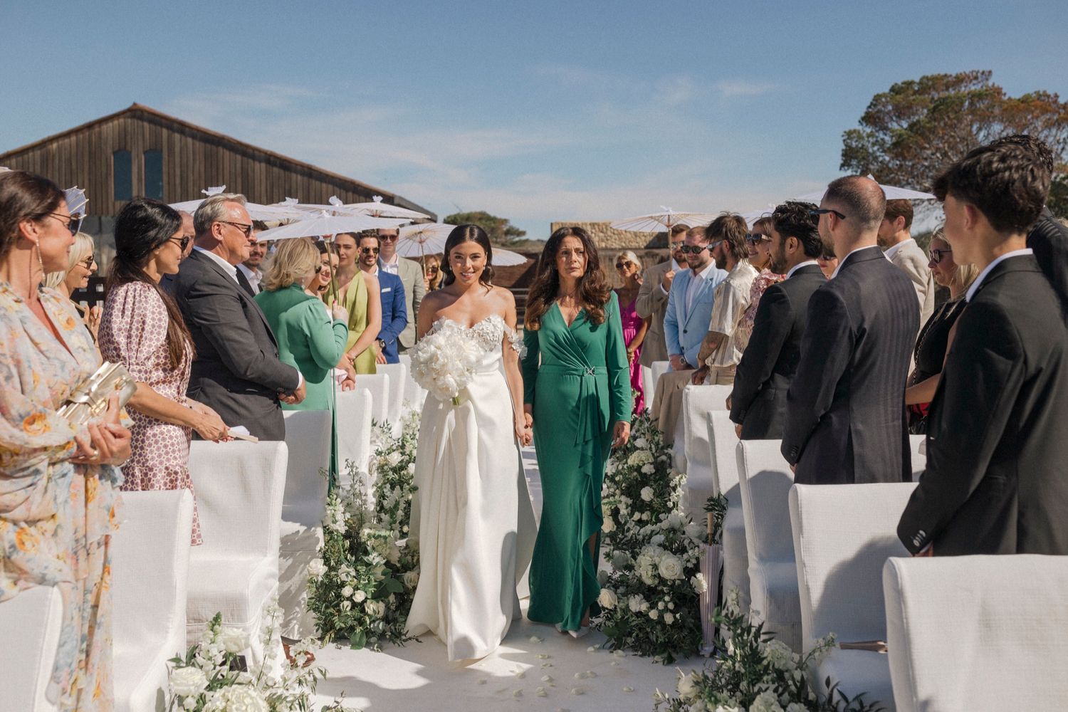 Bride walks down outdoor wedding aisle in white gown, flanked by guests and green floral arrangements under blue sky.