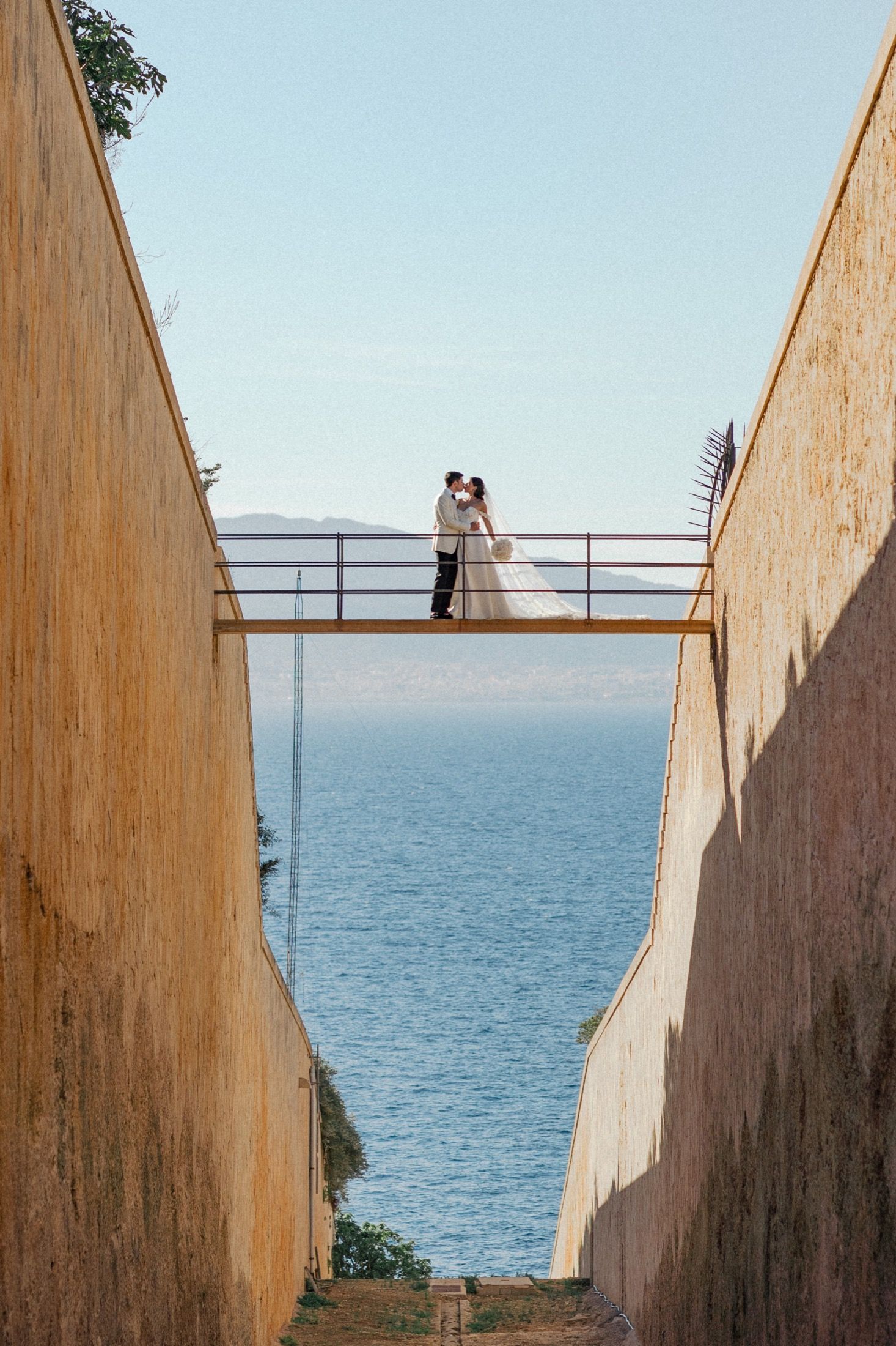 Bride and groom kissing on a bridge between stone walls with blue ocean backdrop on their wedding day.