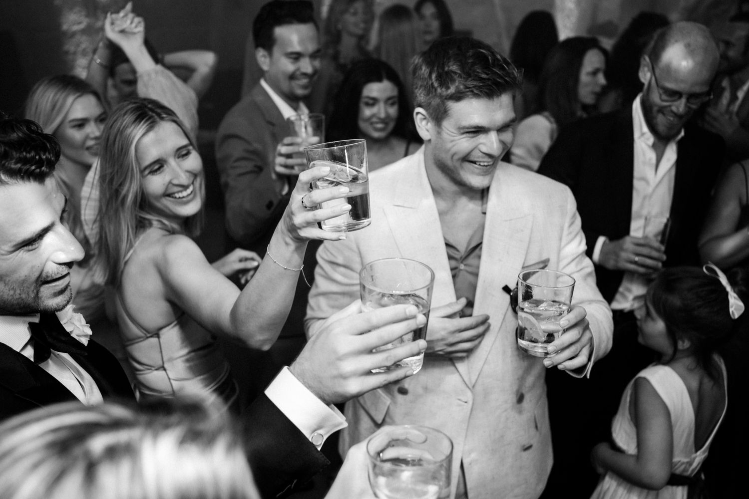 Black and white photo of guests laughing and toasting drinks at a lively wedding reception party.