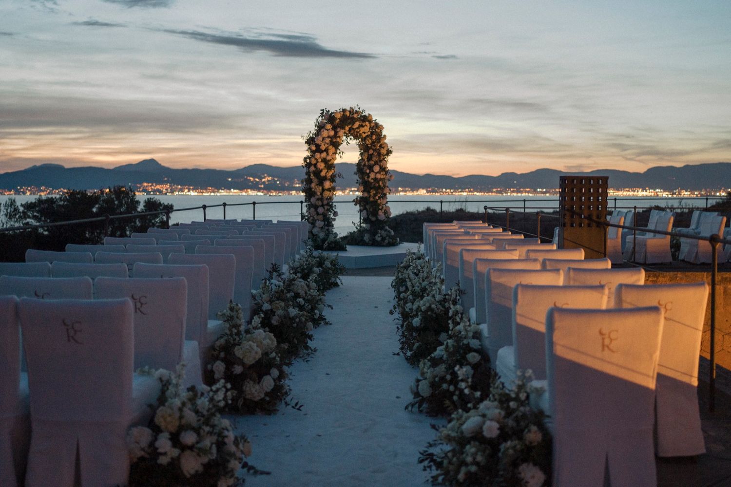 Elegant outdoor wedding ceremony setup at sunset with floral arch, white chairs, and stunning waterfront backdrop.
